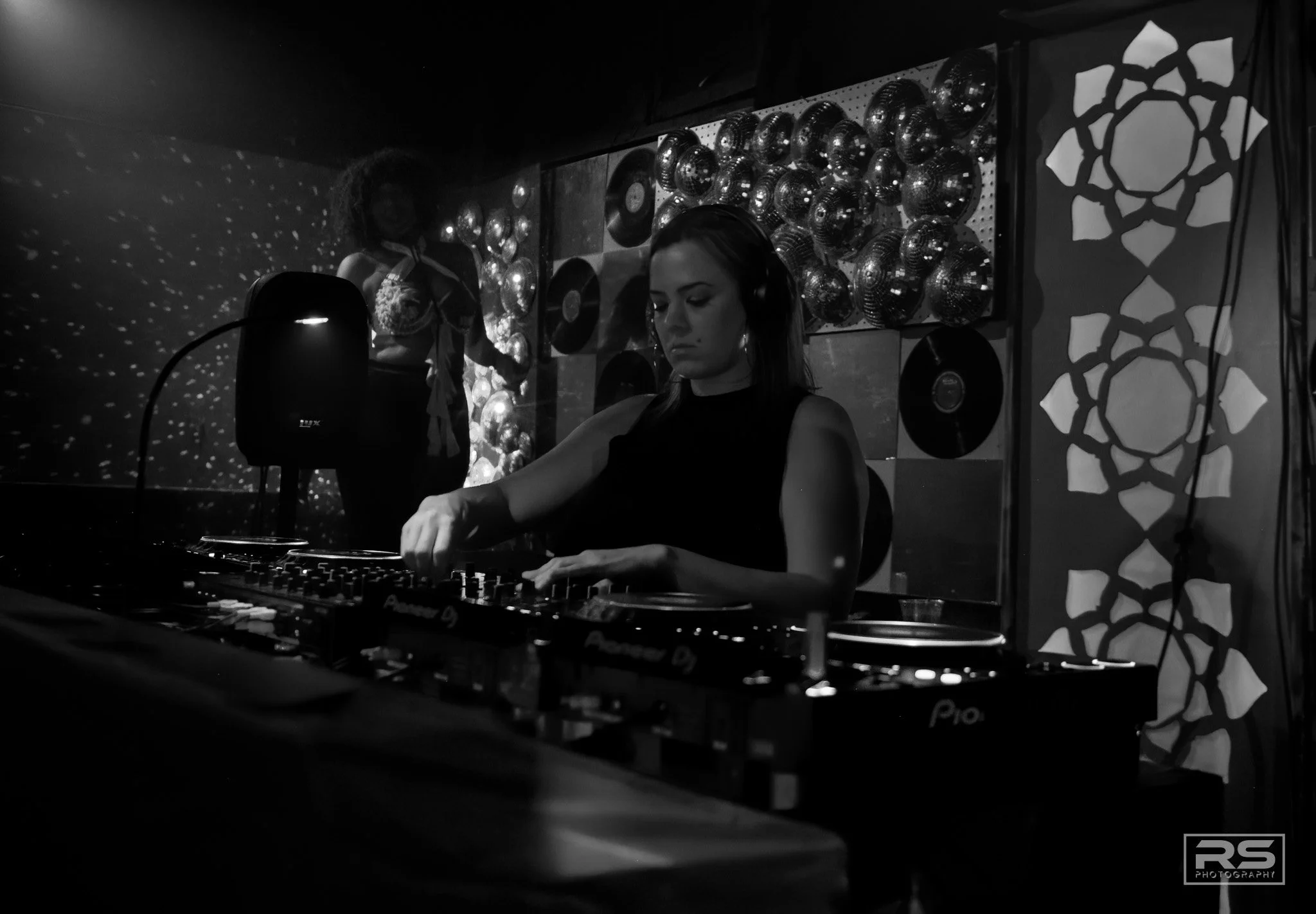A female DJ wearing headphones is playing music at a nightclub with decorative disco balls and patterned walls. The DJ is focused on the equipment, with a dark atmosphere and light reflections on the disco balls.