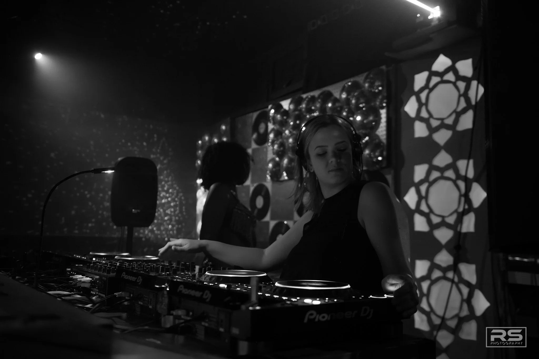 A female DJ wearing headphones is performing at a nightclub with a dark background and decorative lights.