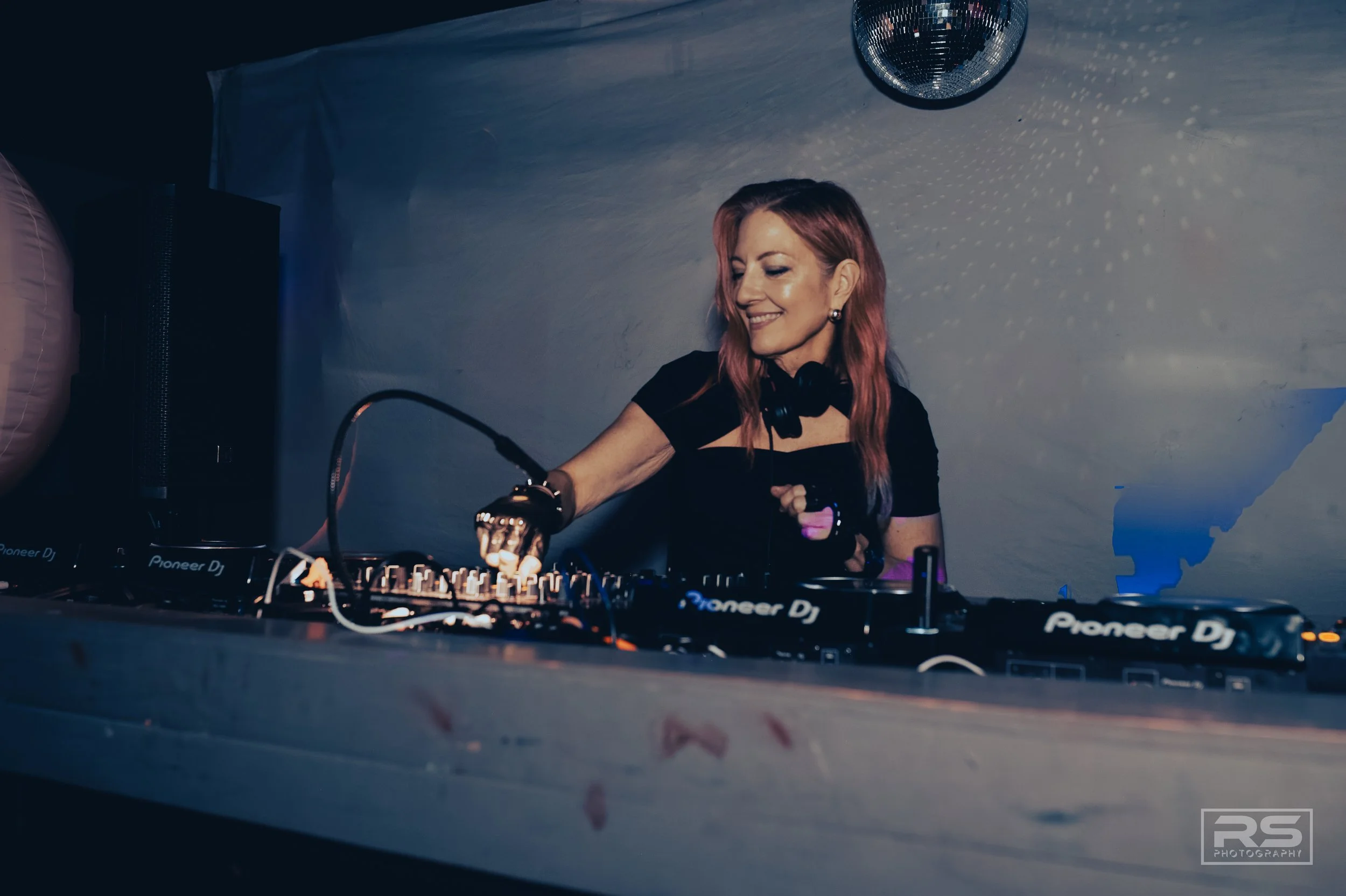A female DJ with pink hair smiling as she mixes music on professional DJ equipment in a dimly lit room with a disco ball overhead.
