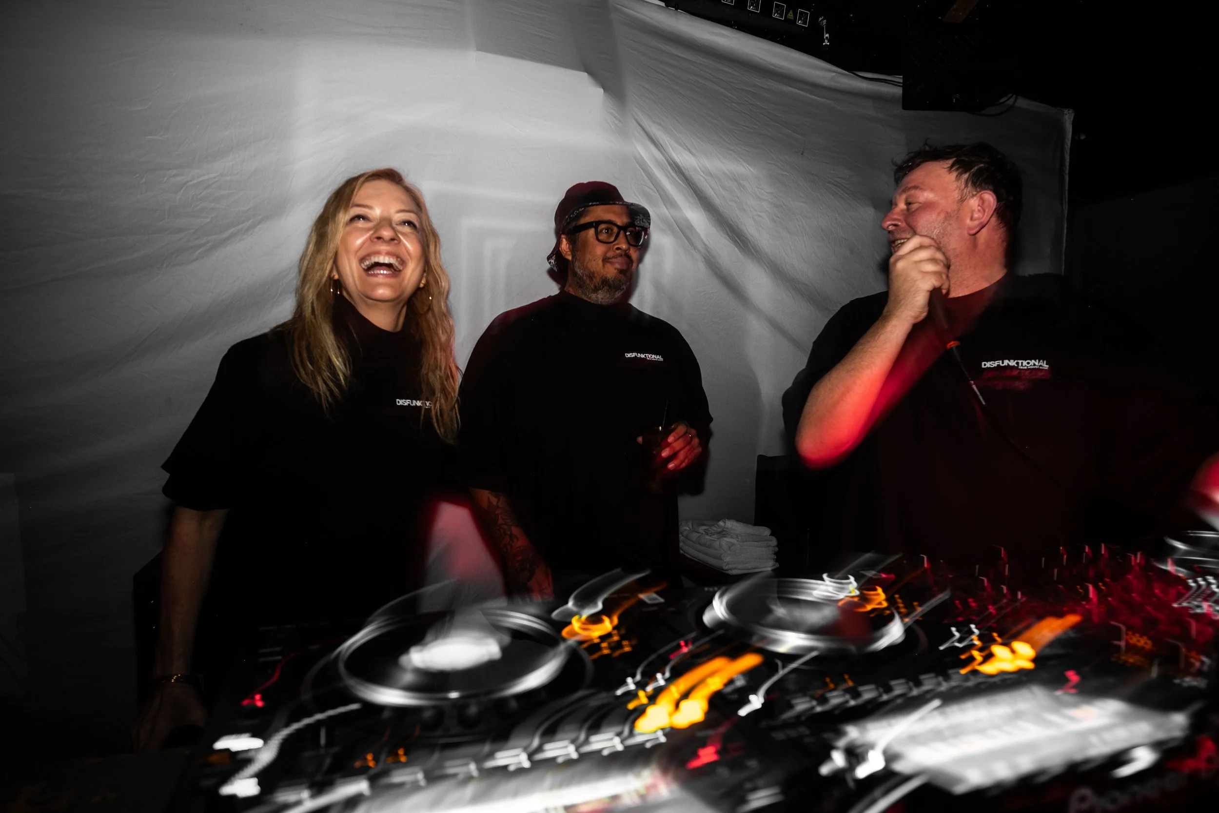 Three people are standing behind a DJ booth, enjoying themselves at a party or event. The woman on the left is smiling broadly, the man in the middle is wearing glasses and a cap, and the man on the right is laughing, with his hand near his face. They are all wearing black shirts with the word 'DISFUNKTIONAL' printed on them. The background is plain, and the DJ equipment is visible in the foreground with colorful, swirling lights.