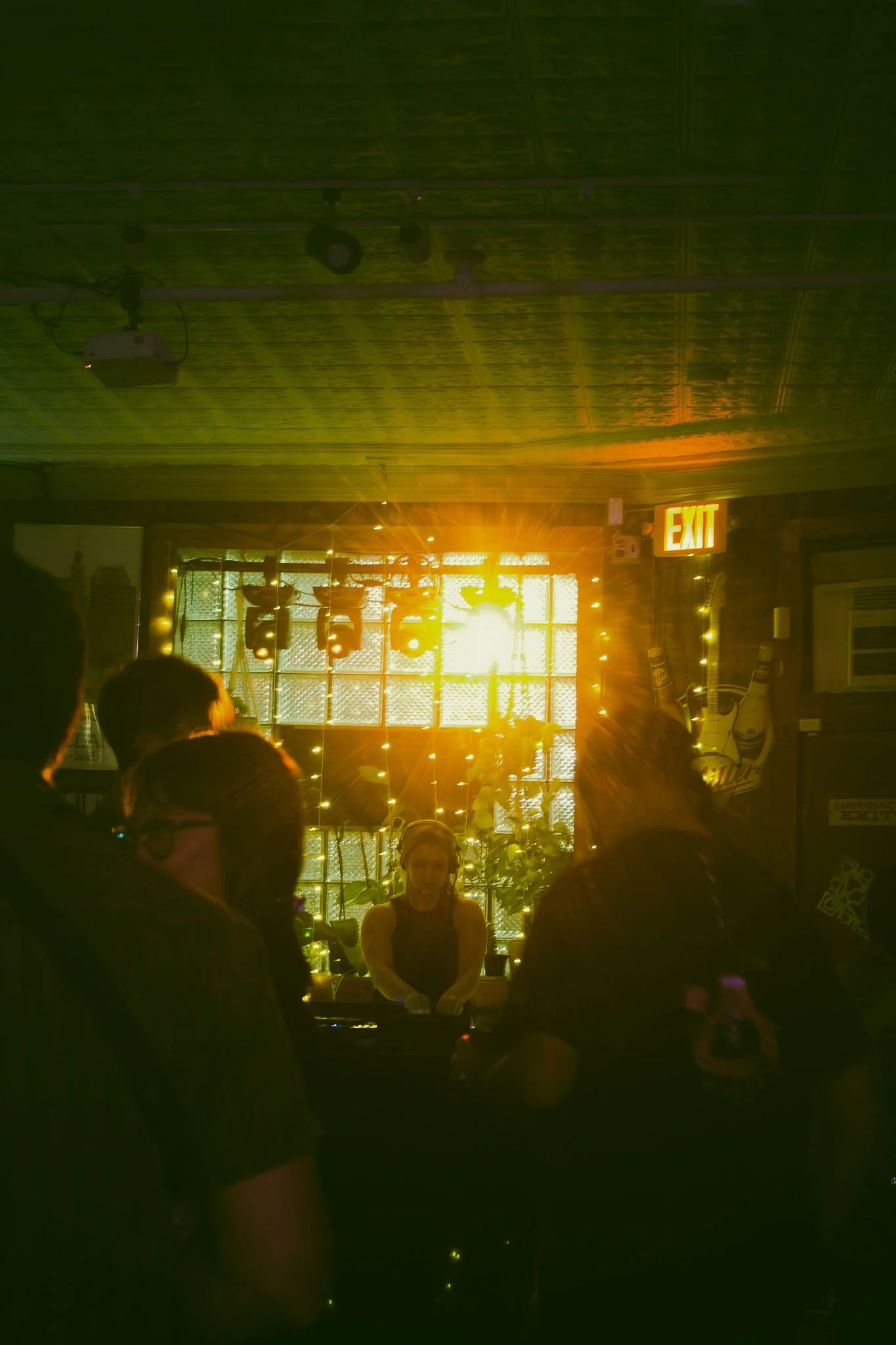 Indoor scene with a stage illuminated by bright yellow sunlight, a person working at a DJ booth, surrounded by people, with string lights and an exit sign visible on the right.
