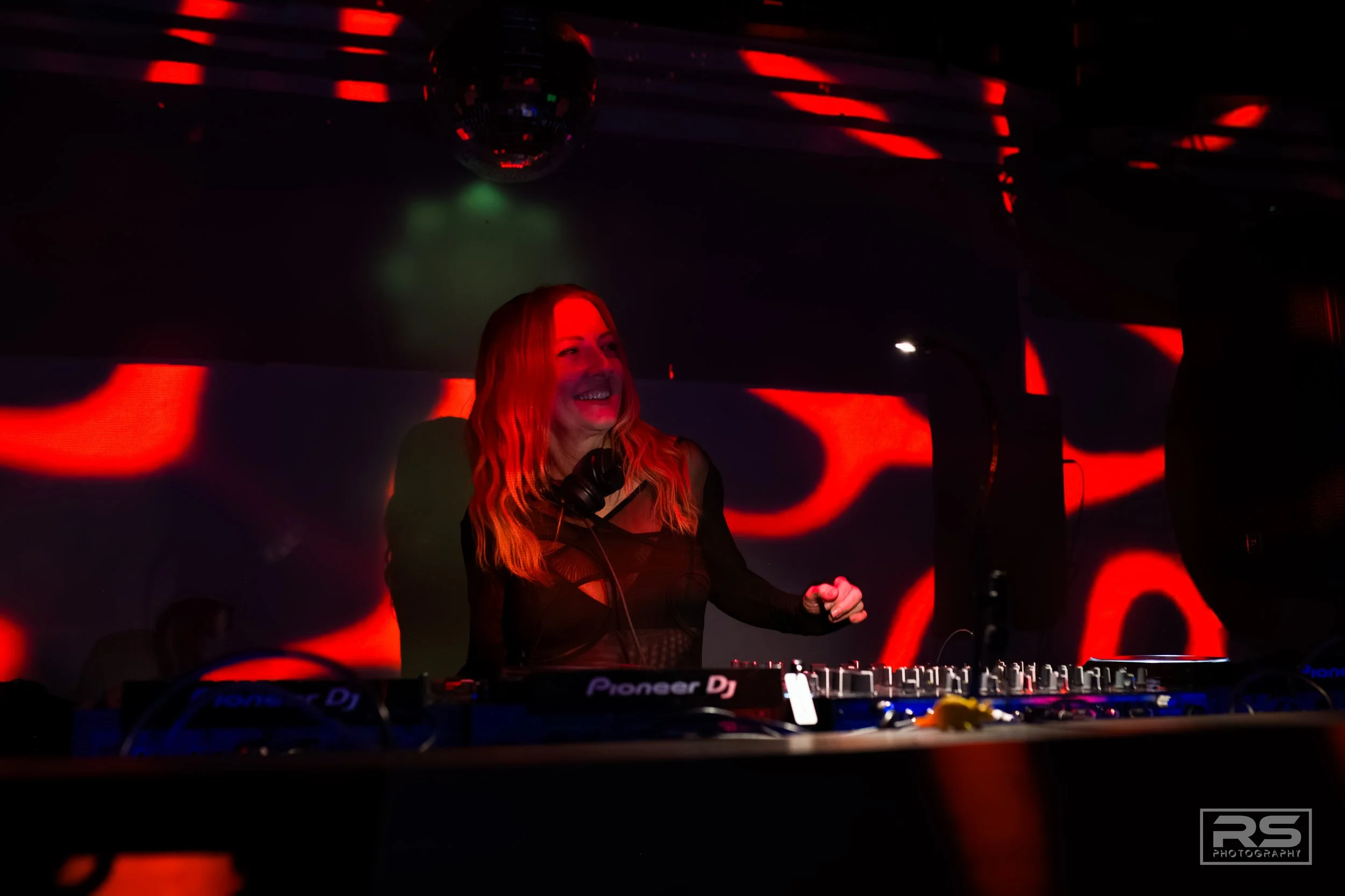 A female DJ with long red hair is performing in a dark club with red and black lighting, standing behind a Pioneer DJ setup.