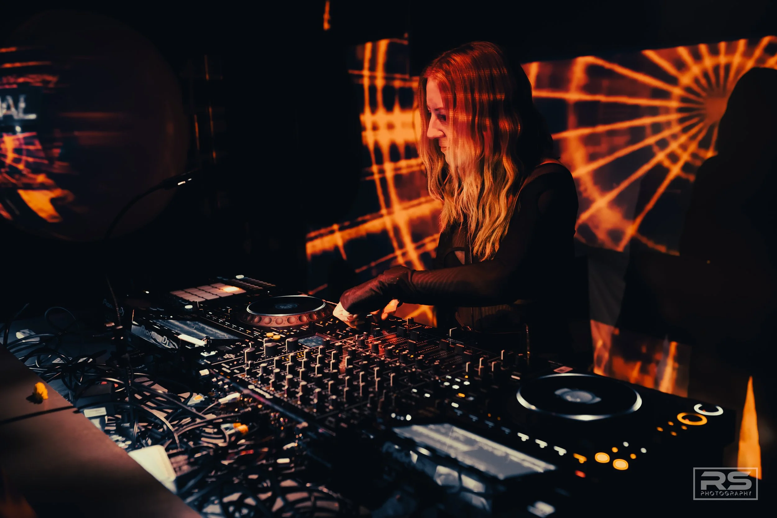 A female DJ with long wavy hair performing with DJ equipment in front of an illuminated backdrop with orange light patterns.