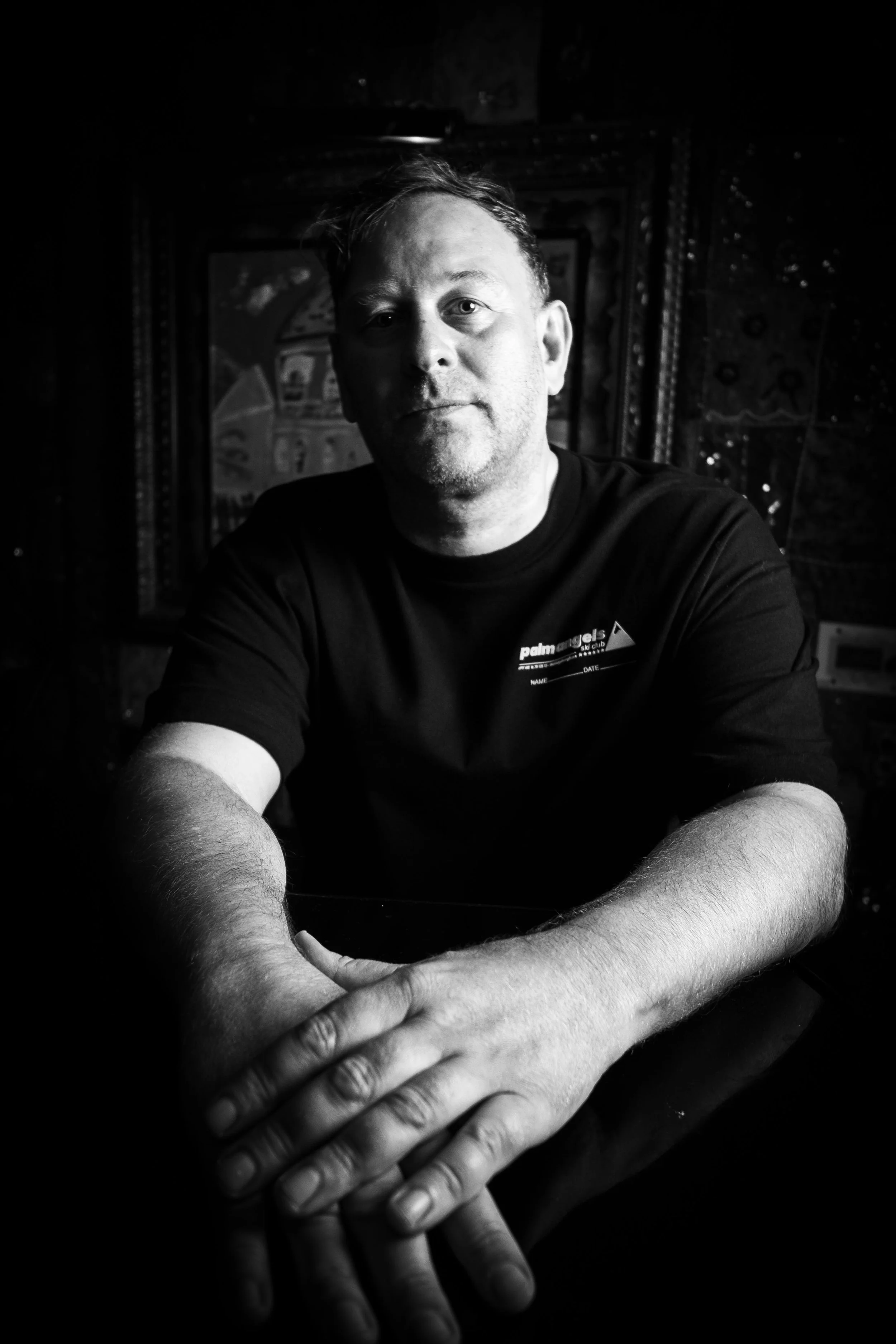 Black and white photo of a man with short hair, sitting at a table with his arms crossed in front of him, and multiple hands reaching towards him from the bottom of the image.