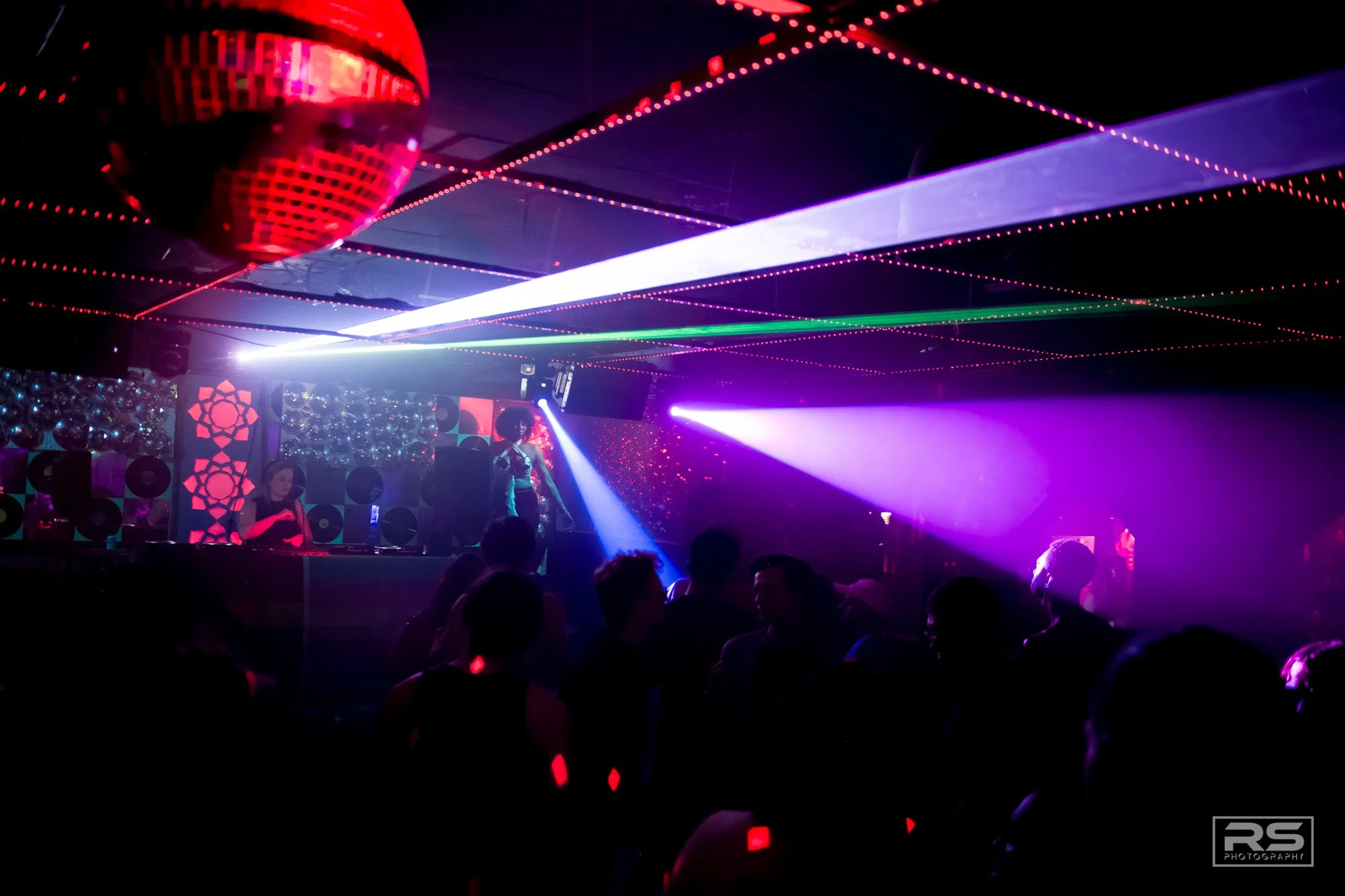 A nightclub scene with laser lights and a DJ performing on stage, with a crowd of people dancing.