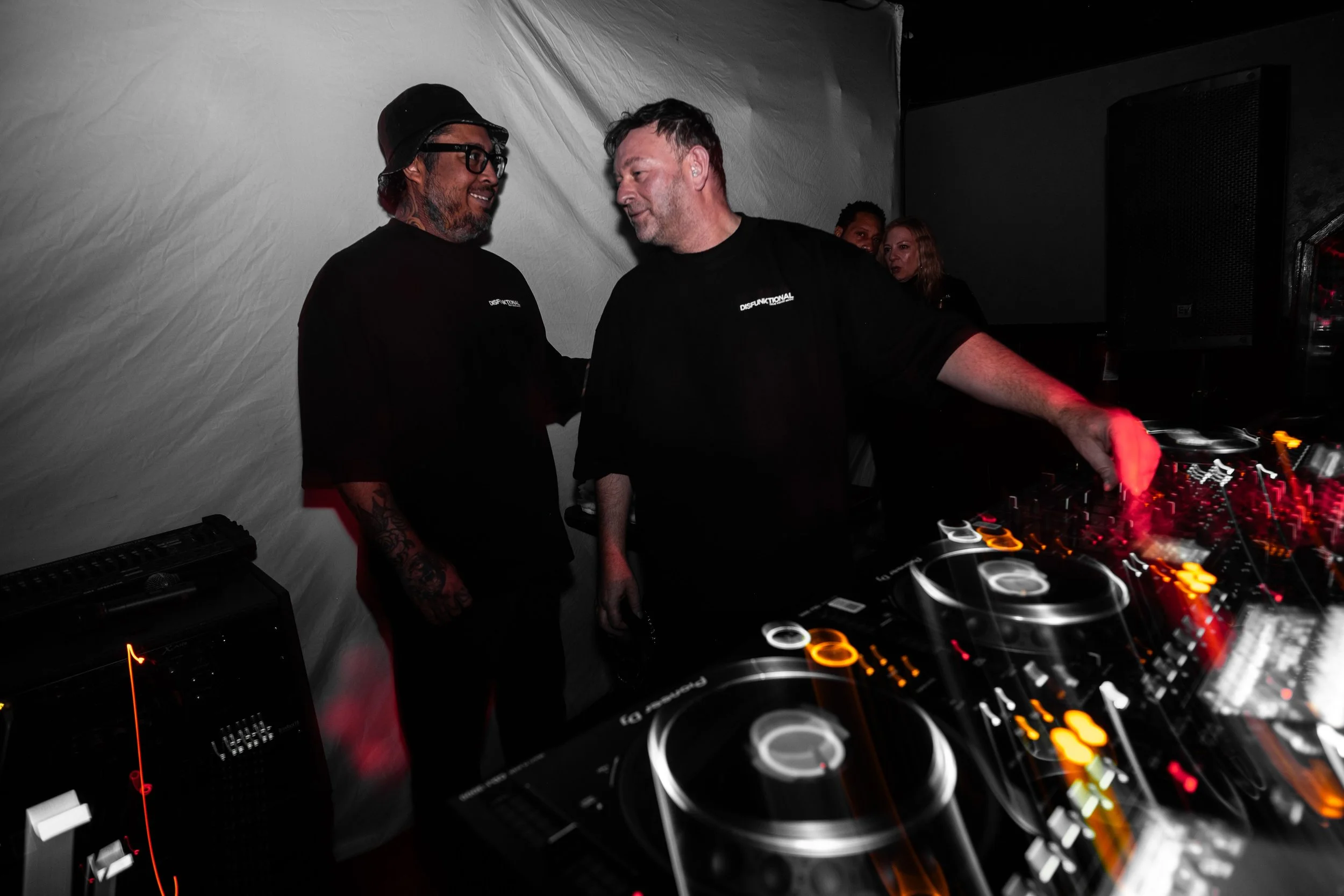 Two men in black shirts with 'DISFUNTIONAL' logos engage in conversation at a DJ booth in a dark room, with a woman and another man in the background, and DJ equipment with glowing controls visible in the foreground.