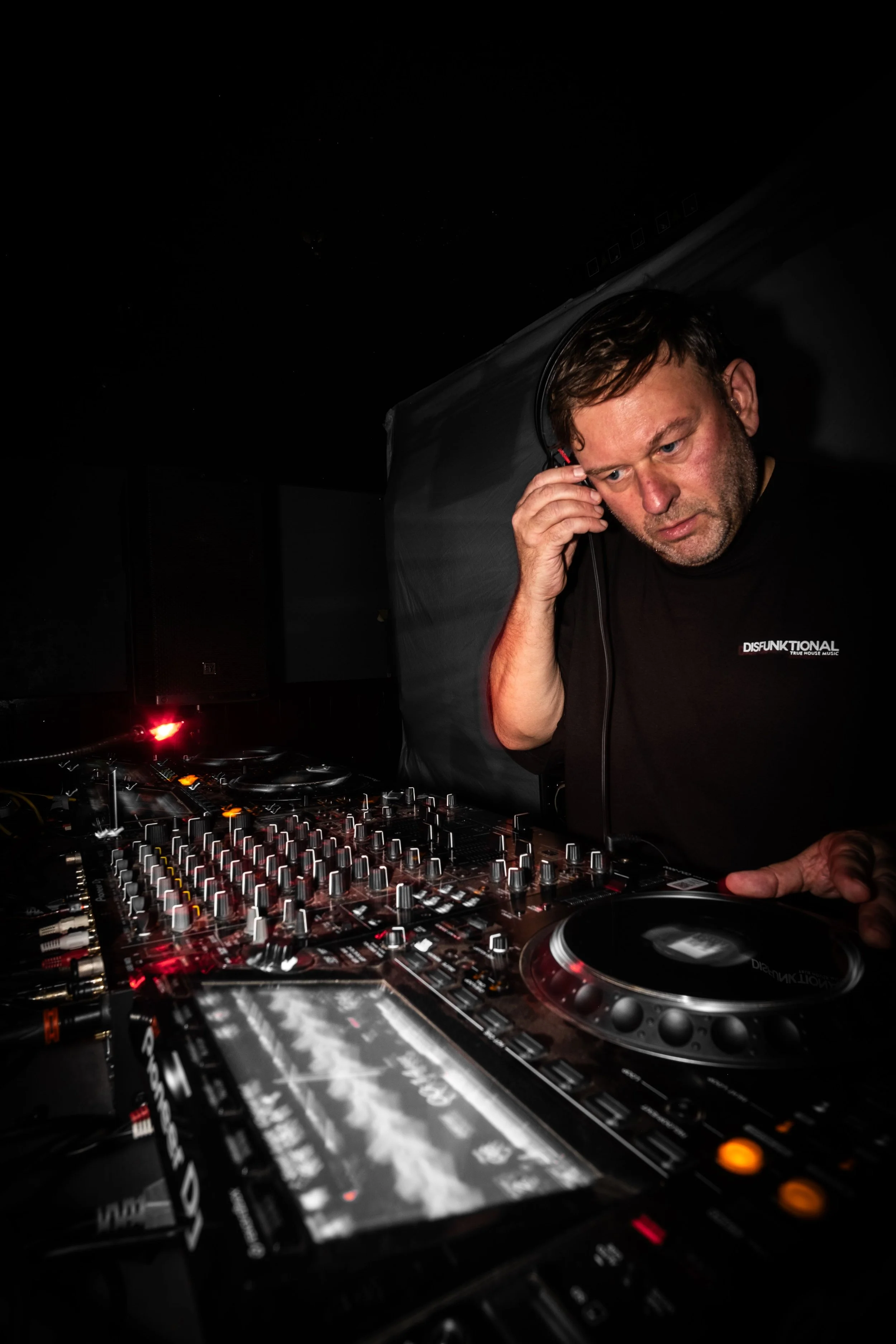 A DJ with headphones adjusting sound equipment in a dark club environment.