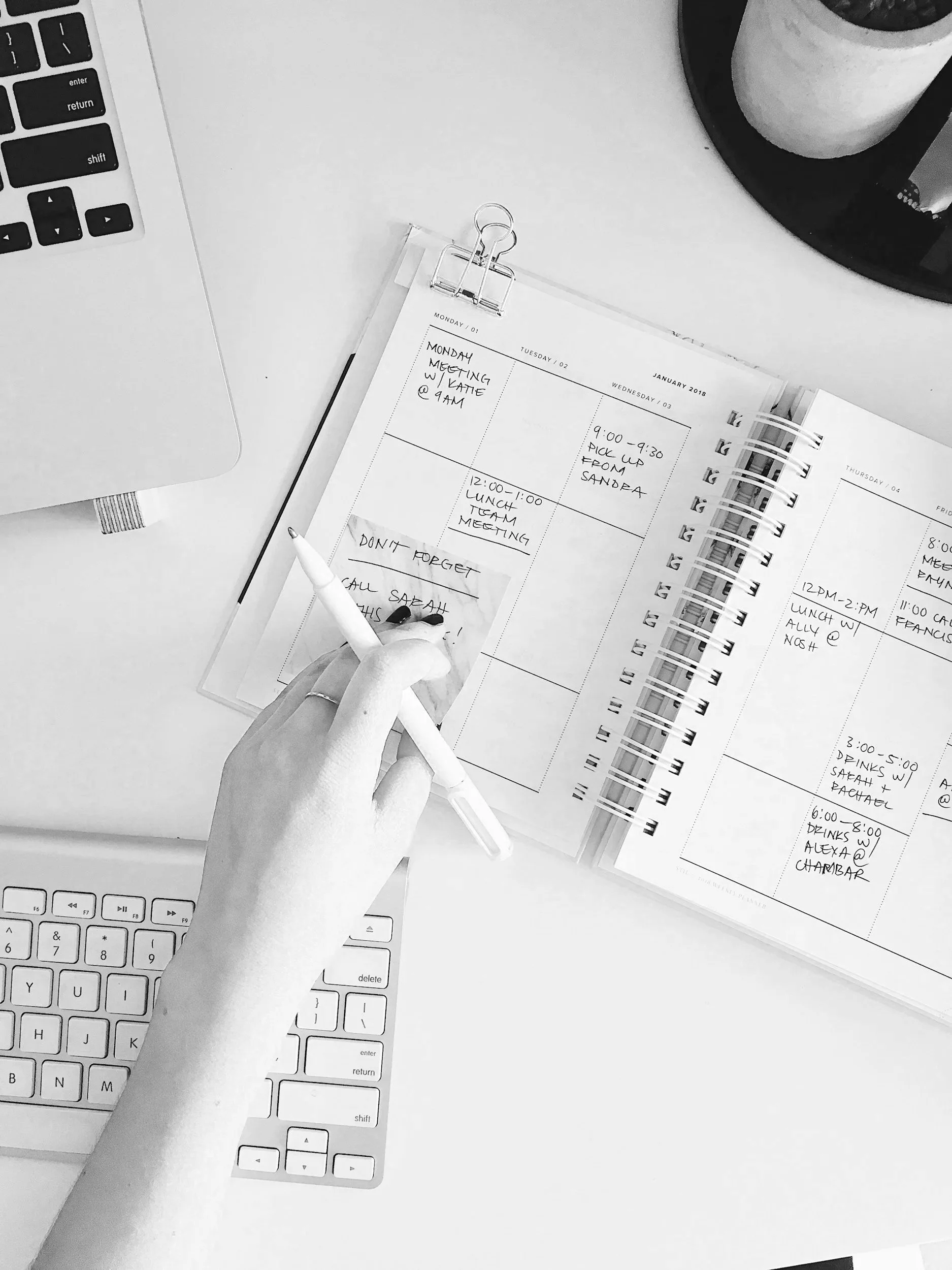 A person writing in a planner with a white pen, surrounded by a laptop, a plant, and a tray on a white desk.