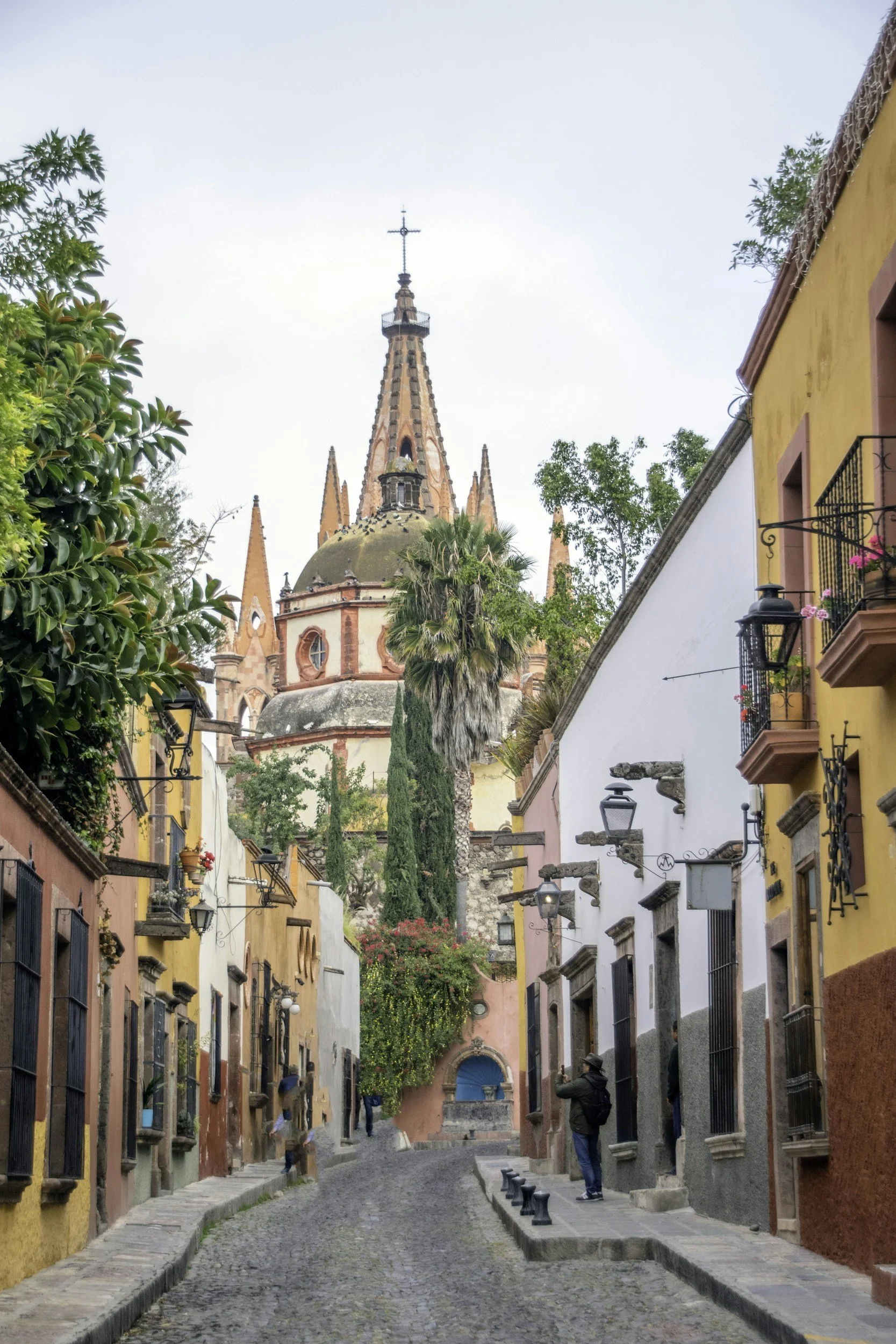 A cobblestone street lined with colorful buildings, with a church tower in the background, lush trees, and people walking and taking pictures.