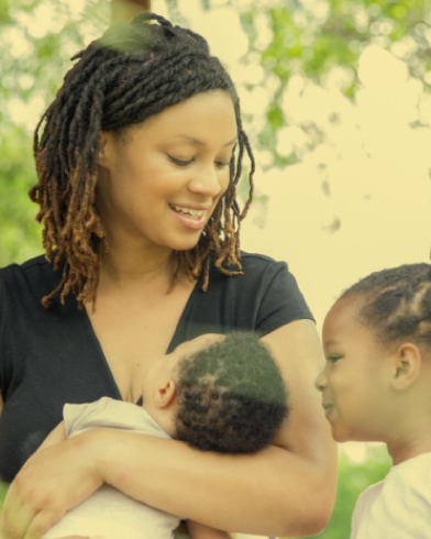 Woman holding a baby and smiling at a young girl outdoors.