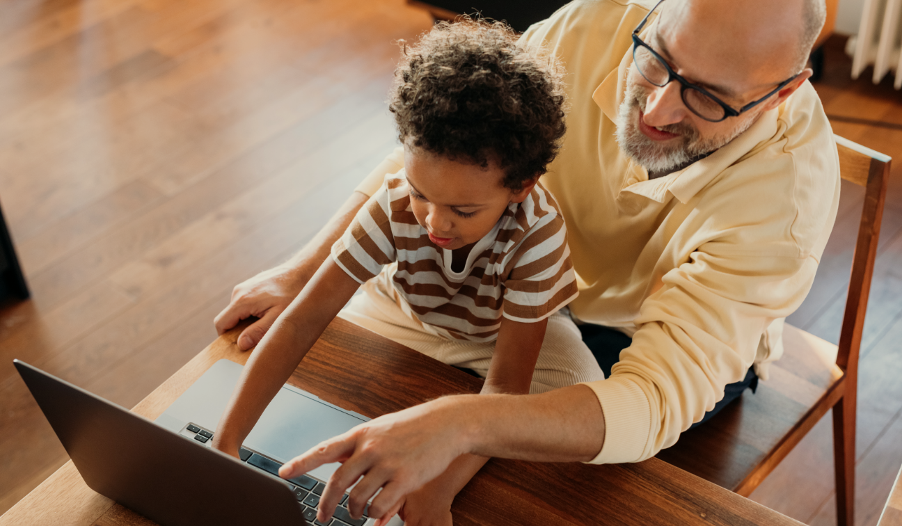 An older man with glasses and a beard teaches a young boy with curly hair how to use a laptop at a wooden table.