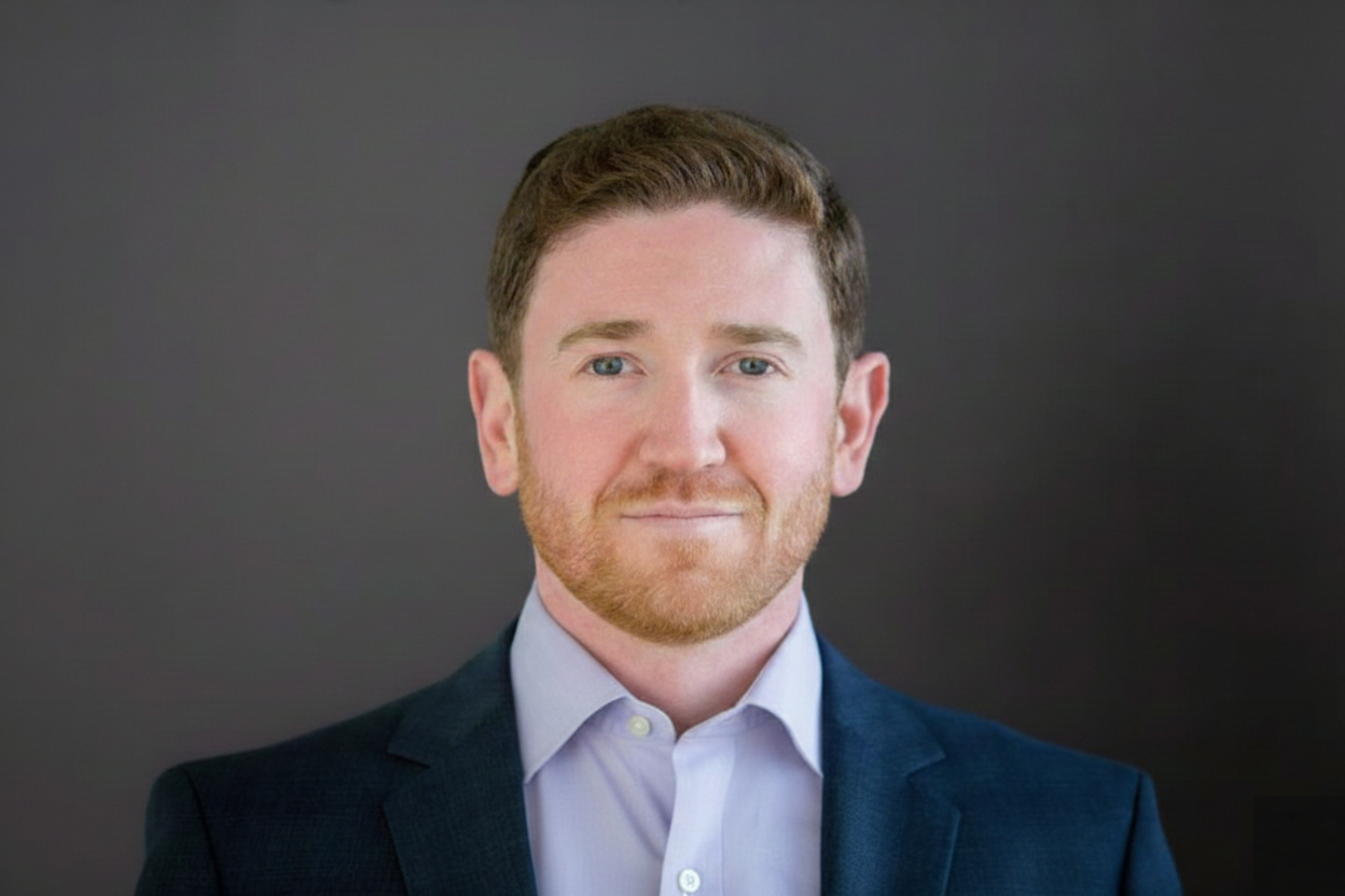 A man in a navy blazer and light blue open collared dress shirt against a dark gradient background, looking directly at the camera with a slight smile.