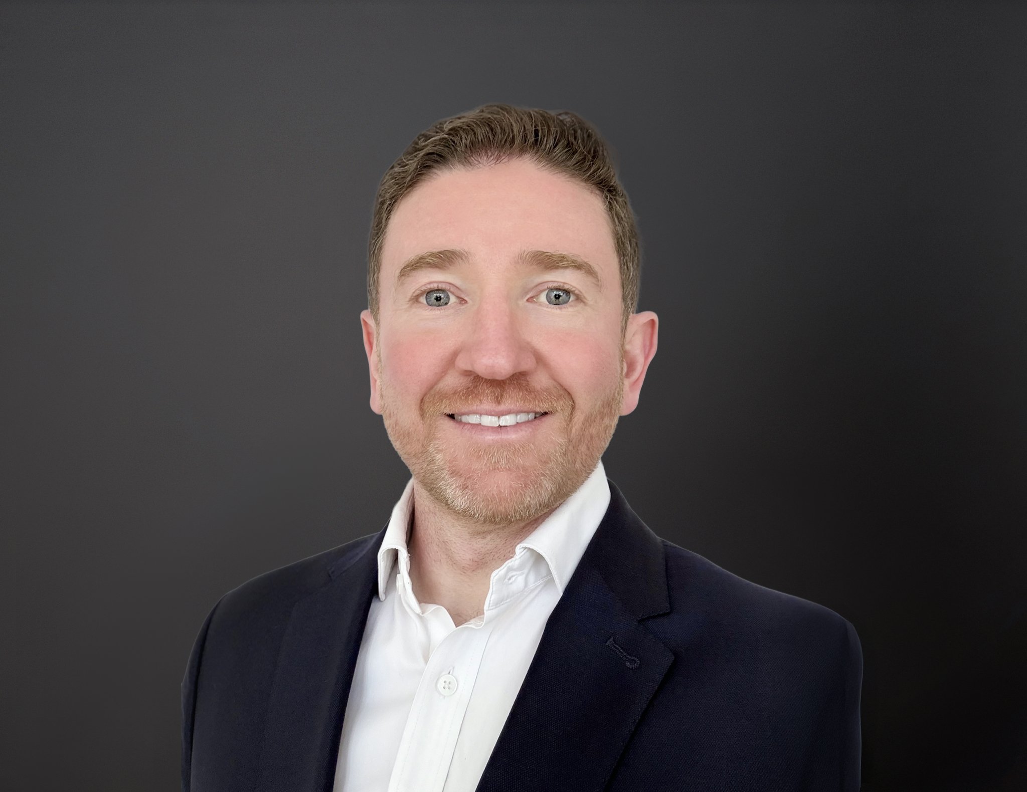 Headshot of a smiling man with short brown hair, a beard, light complexion, wearing a dark blazer and a white shirt, against a dark gray background.