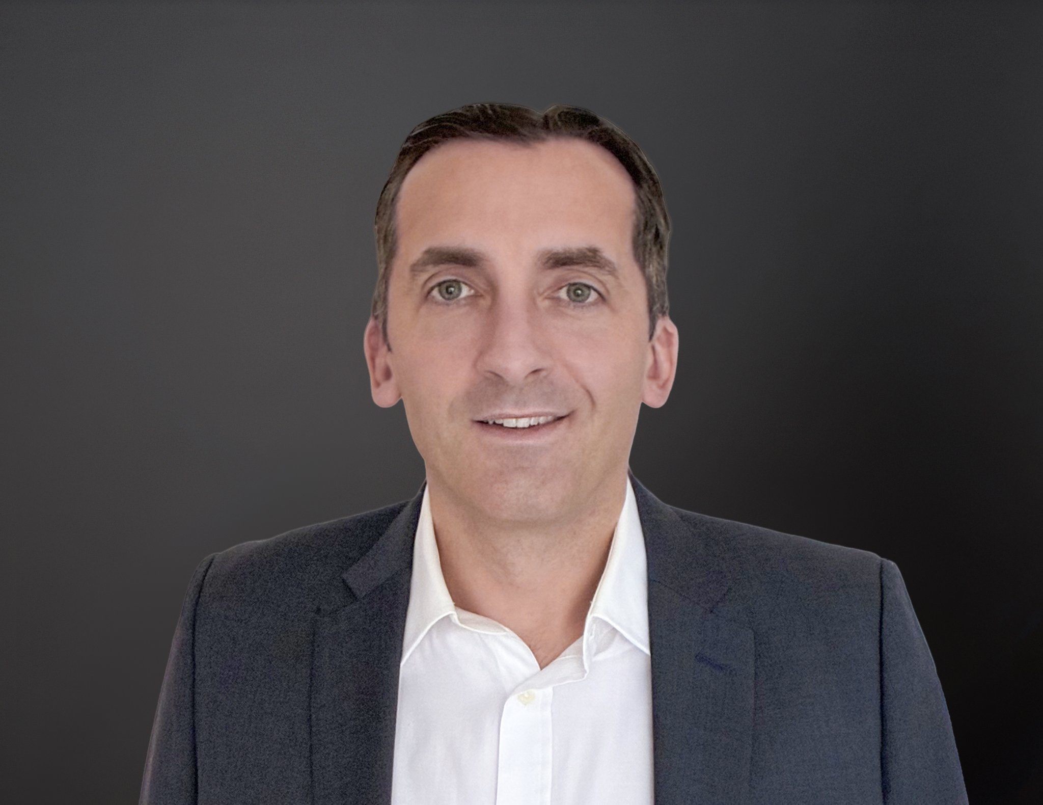 Portrait of a man with dark hair wearing a dark suit jacket and a white shirt, smiling against a dark background.