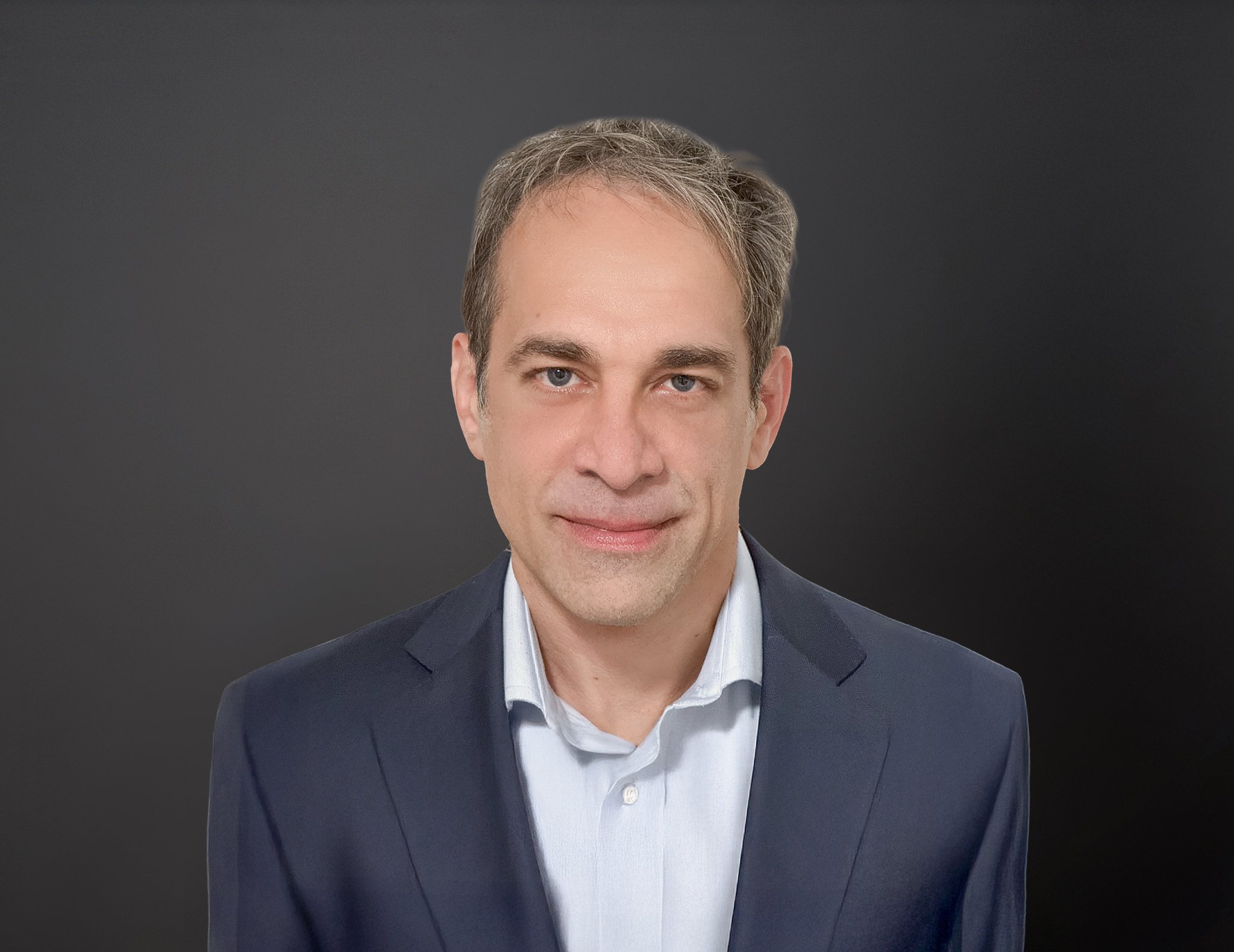 A man in a navy blazer and white shirt against a dark gradient background, looking directly at the camera with a slight smile.