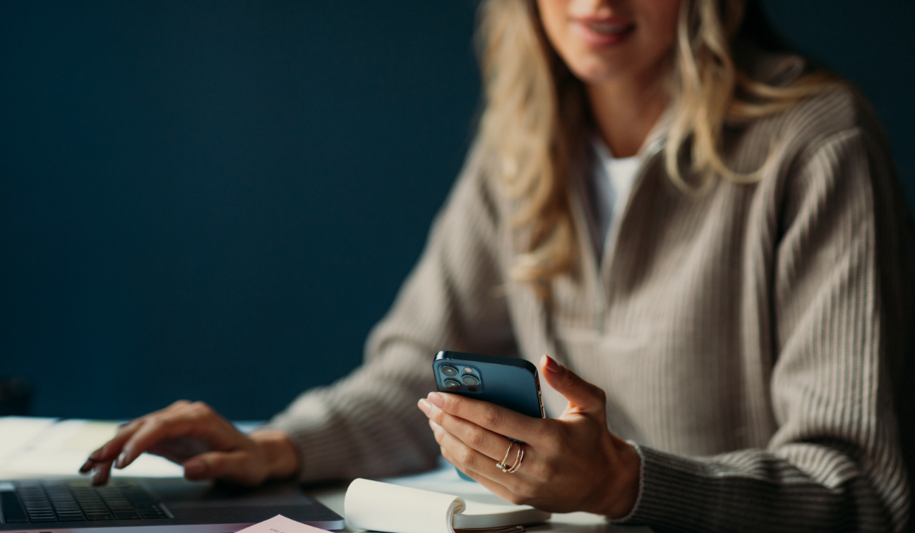 A woman sitting at a desk holding a smartphone and looking at it, with a laptop and notepad on the desk.