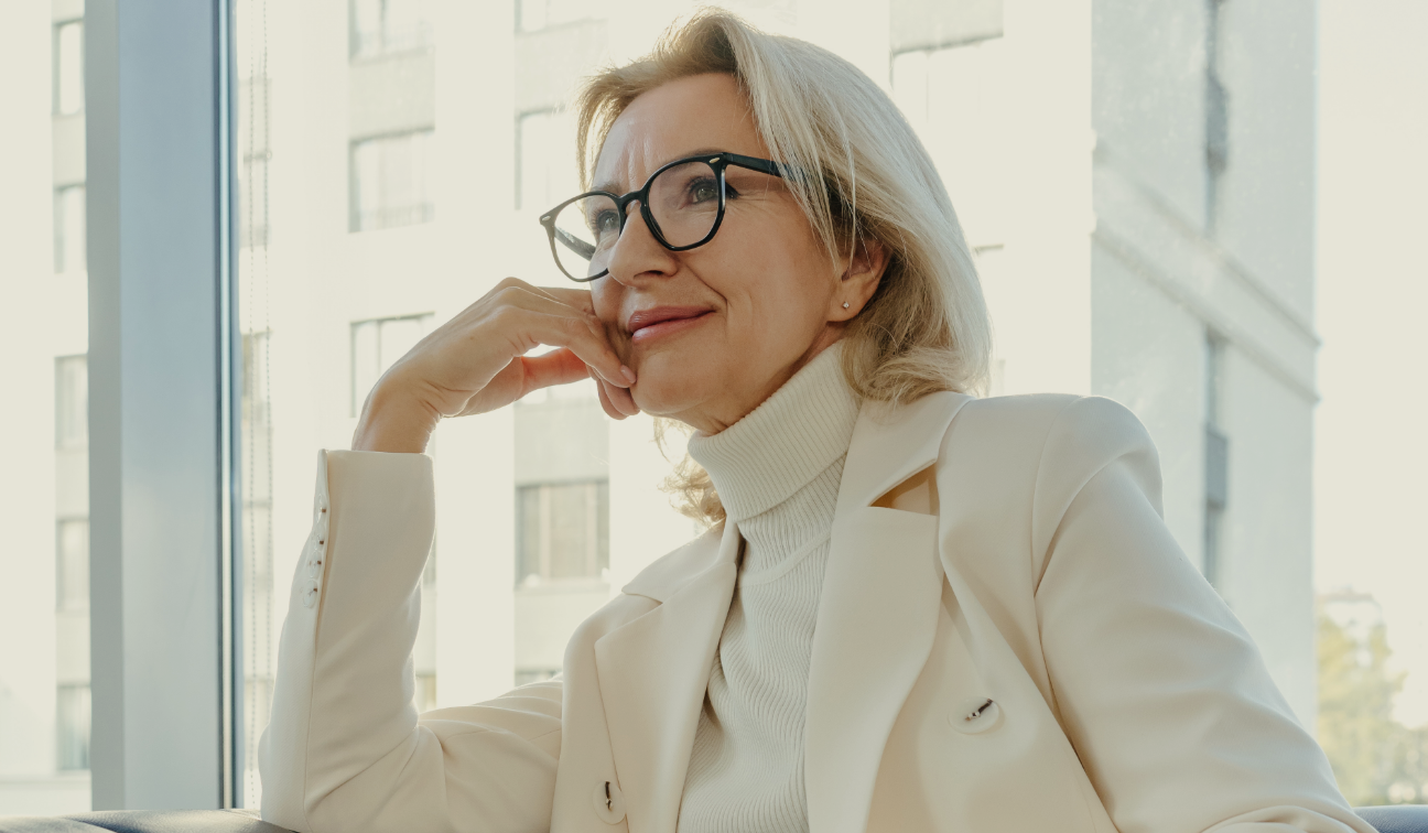 A middle-aged woman with blonde hair and glasses sitting near a window, smiling thoughtfully, wearing a cream-colored blazer and turtleneck.