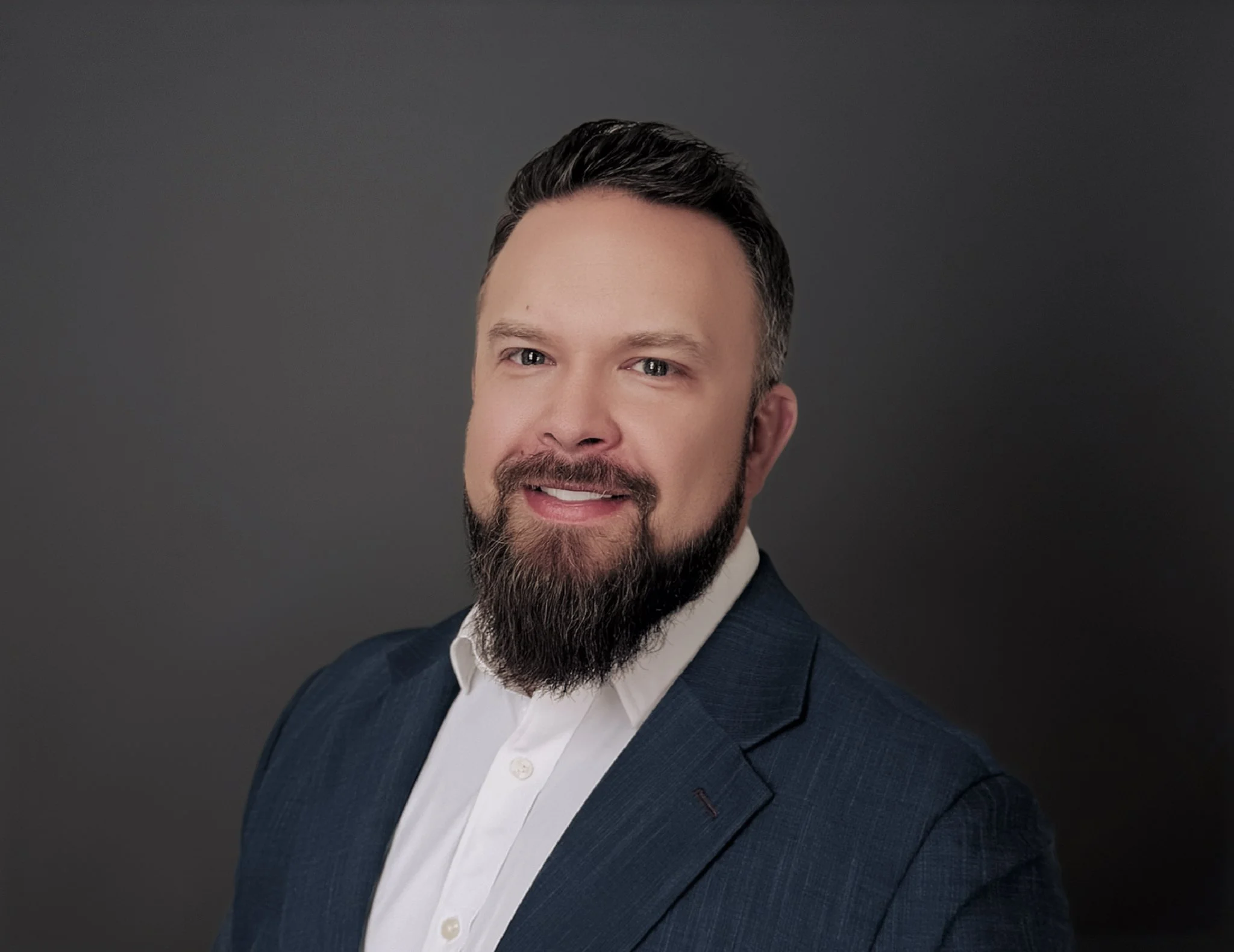 Portrait of a man with a beard and mustache wearing a white shirt and dark blazer against a dark background.