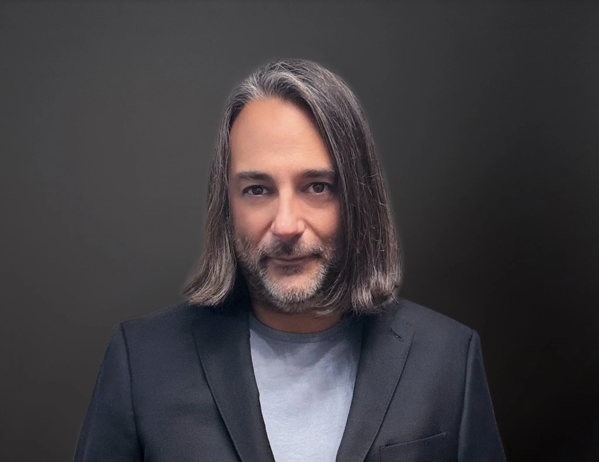 Man with shoulder-length gray hair and a beard, wearing a dark blazer over a light gray T-shirt, against a dark gray background.