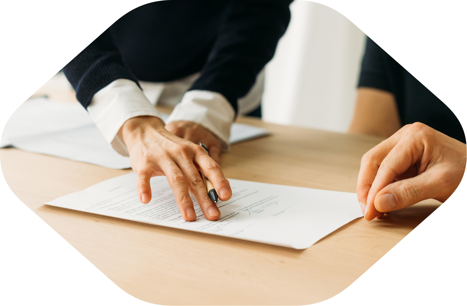 Person signing a document on a wooden table with another person’s hand visible.
