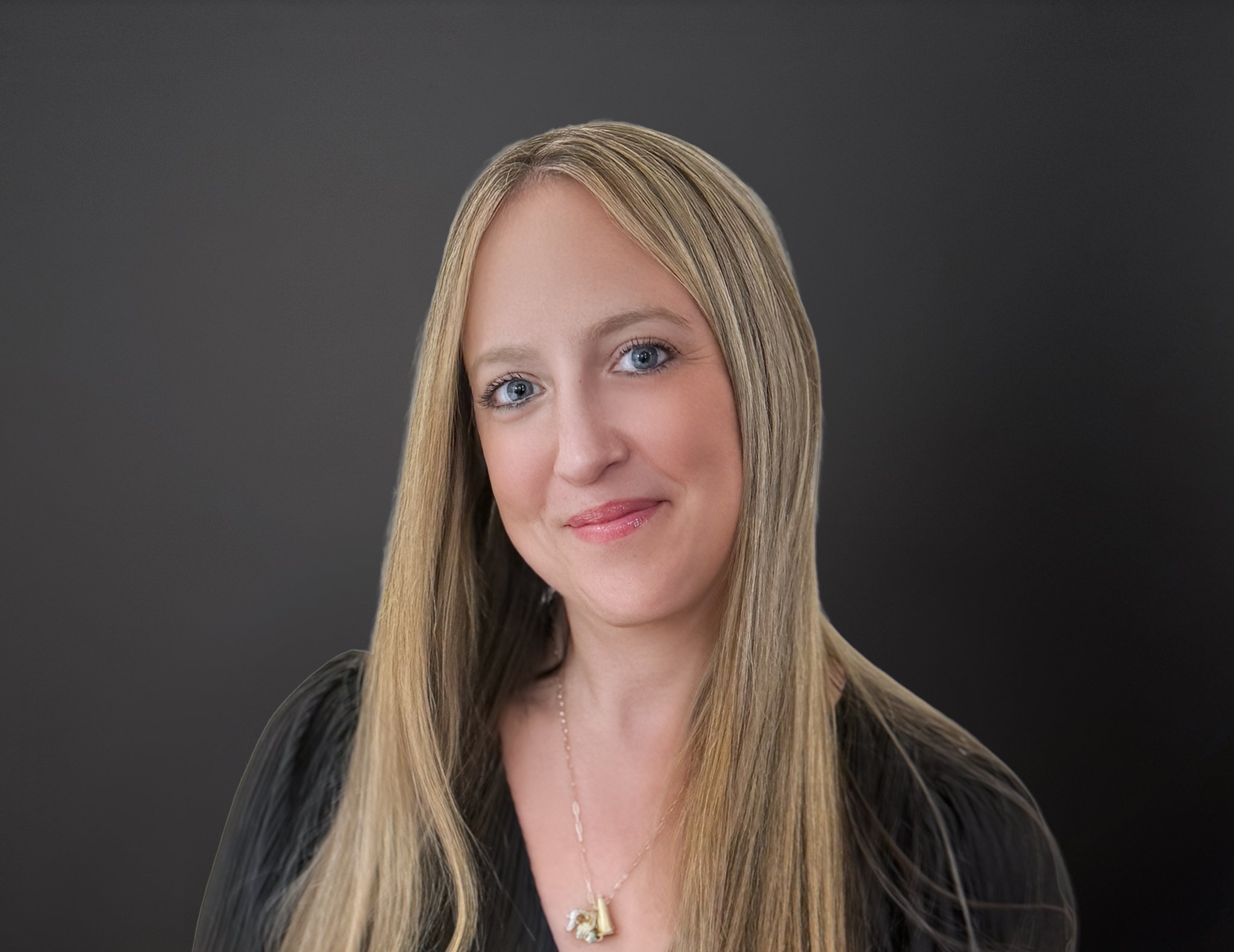 Portrait of a woman with long blonde hair, blue eyes, and a slight smile, wearing a black top and a gold necklace, against a dark gray background.