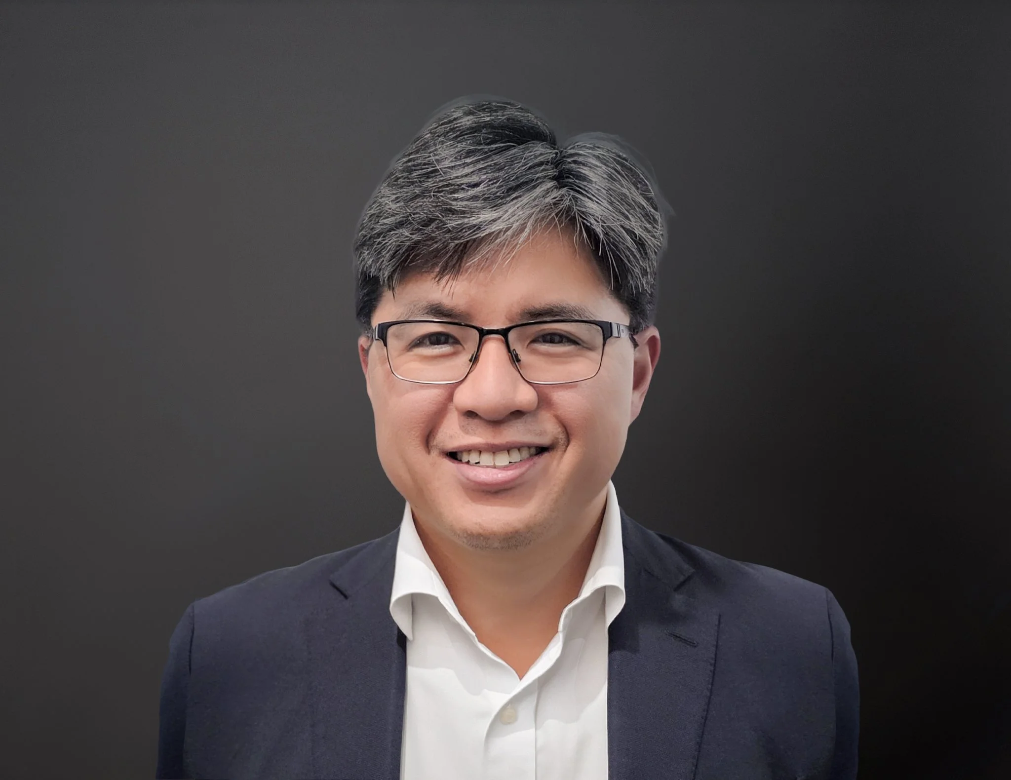 Portrait of a smiling man with glasses, wearing a white shirt and dark blazer, against a dark background.