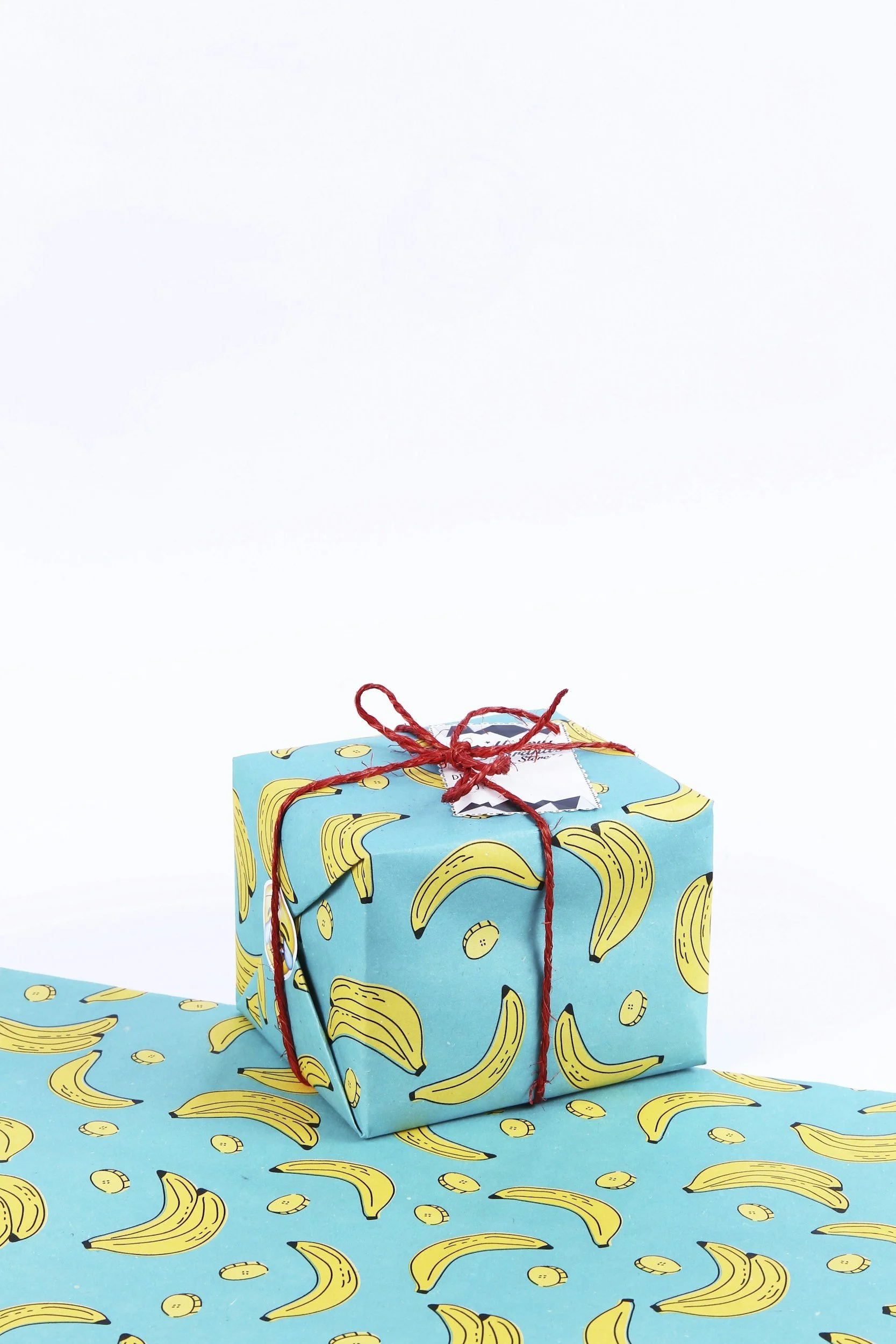 Gift wrapped in blue paper with banana print, tied with red string, placed on matching banana print wrapping paper.