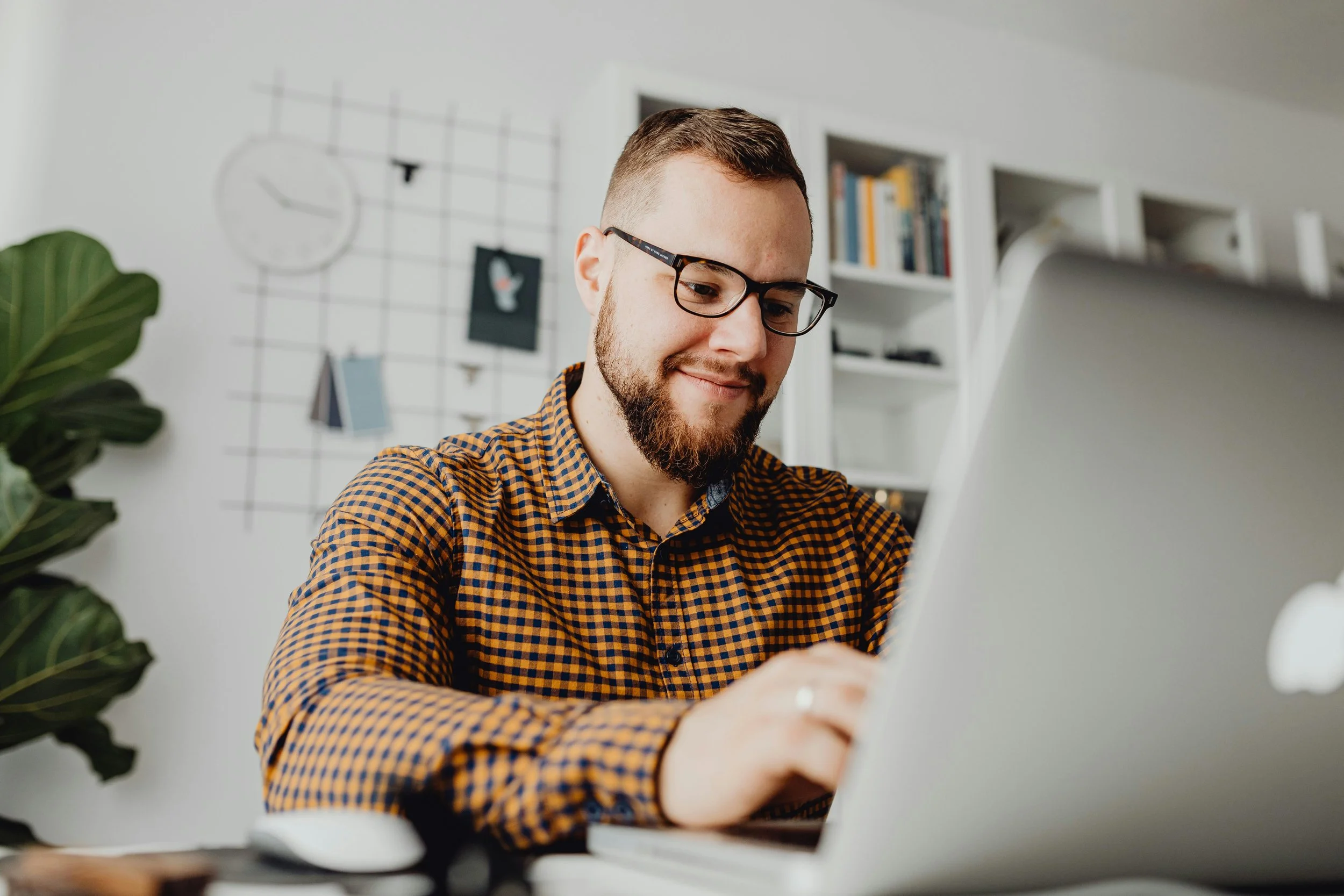 A man with glasses and a beard sitting at a desk using a laptop, with a bookshelf and decorative wall grid in the background.