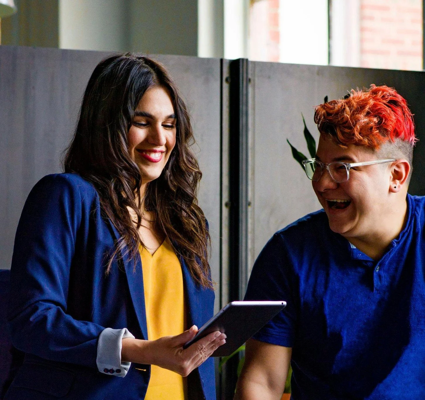 Two people, a woman with long dark hair wearing a yellow shirt and blue blazer, and a man with short red hair, glasses, and a blue shirt, are smiling and looking at a tablet together in an office setting.