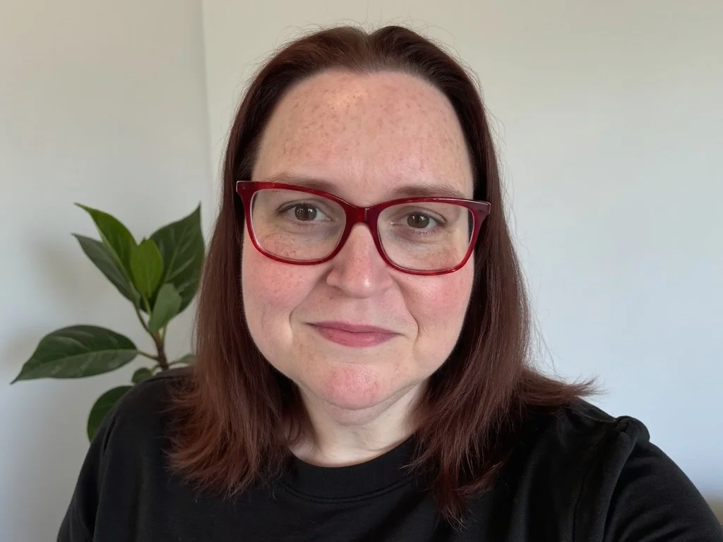A woman with shoulder-length brown hair, wearing glasses with red frames and a black top, smiling at the camera with a green houseplant in the background.