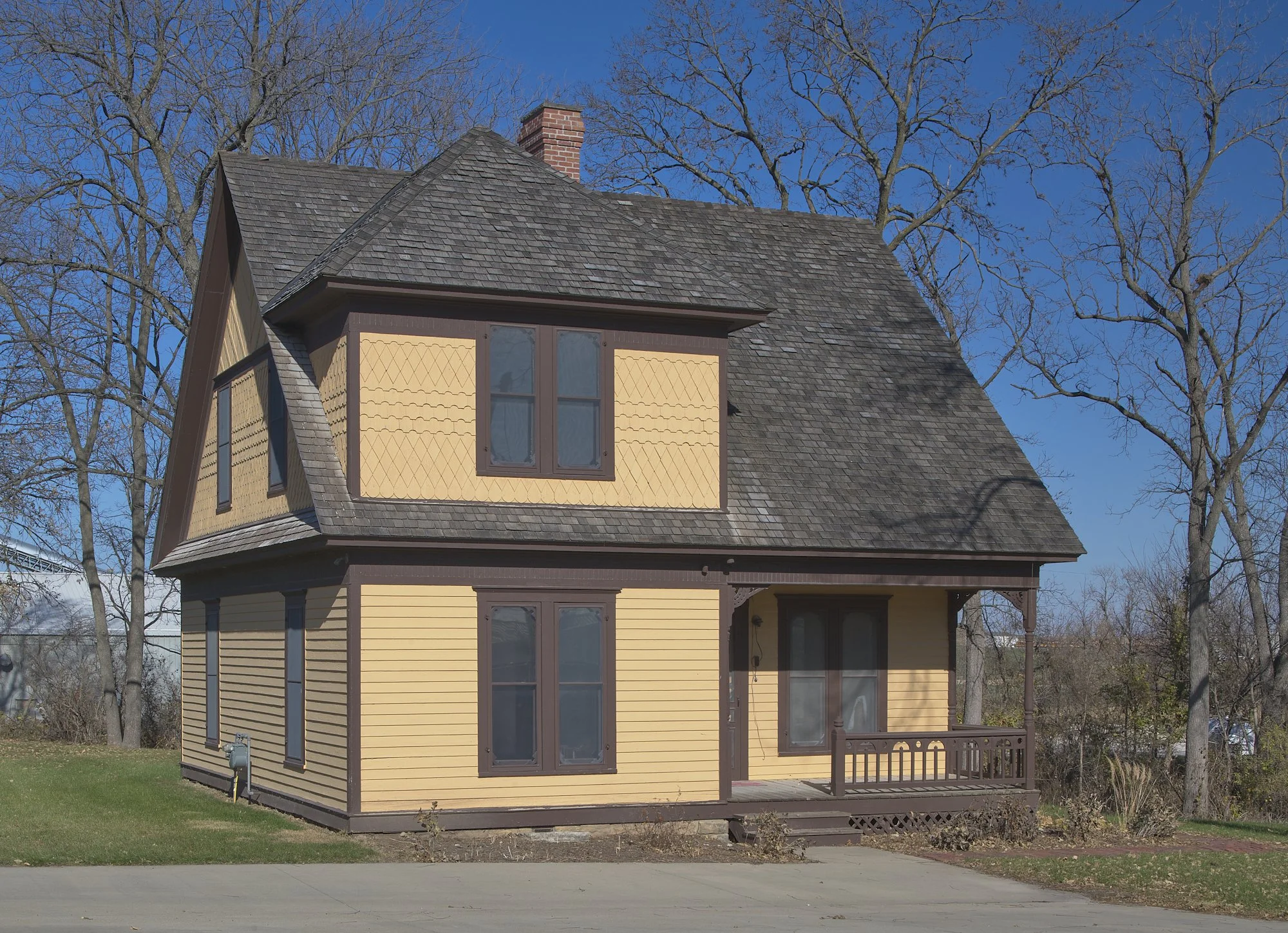 Historic yellow two-story house with brown trim, a steep shingled roof, and front porch surrounded by leafless trees and grass under a clear blue sky.