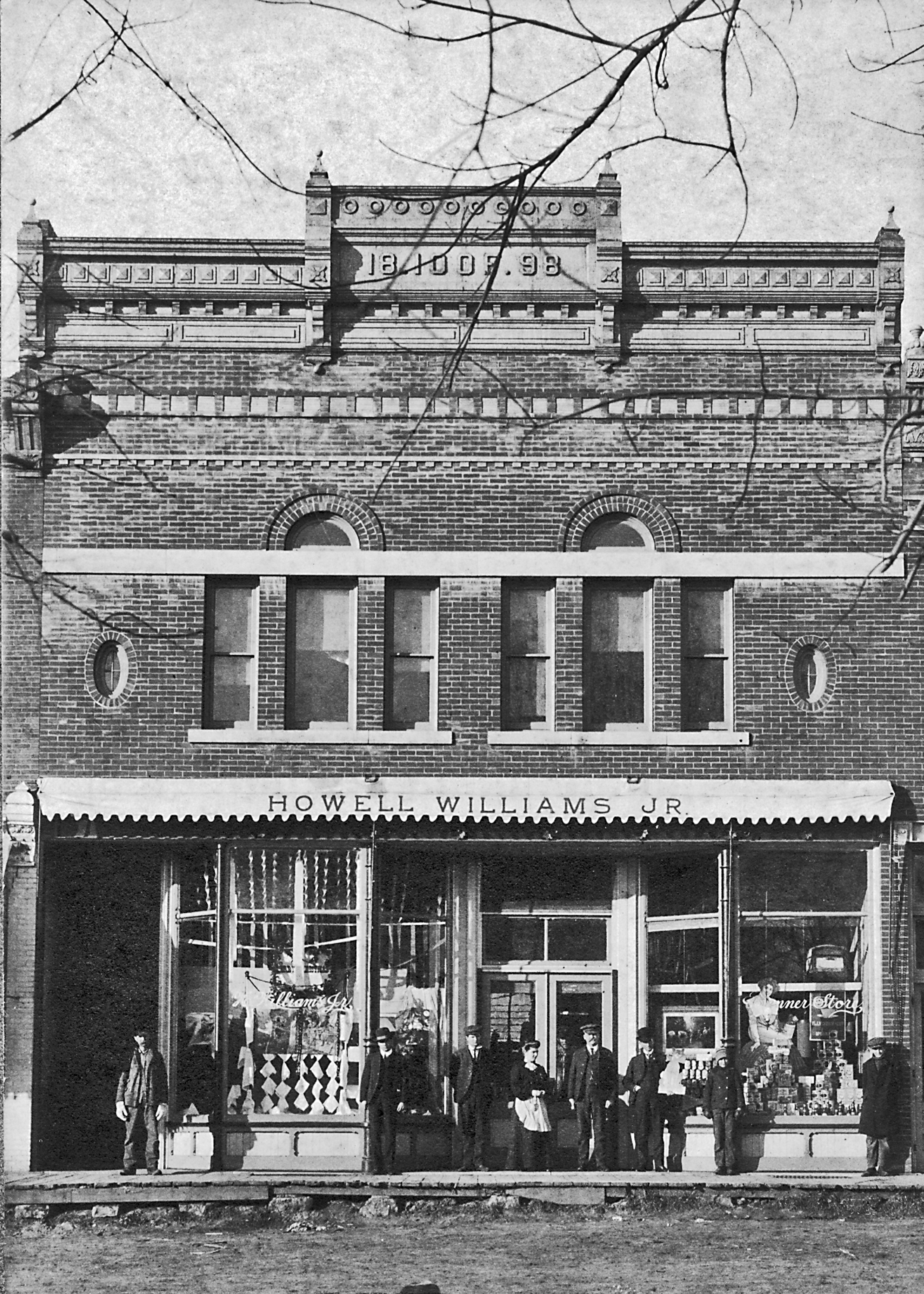 A black-and-white photo of a historic brick storefront with the name 'Howell Williams Jr.' on a sign above the entrance. Several people are standing outside the shop, and the display windows show various items and signs.