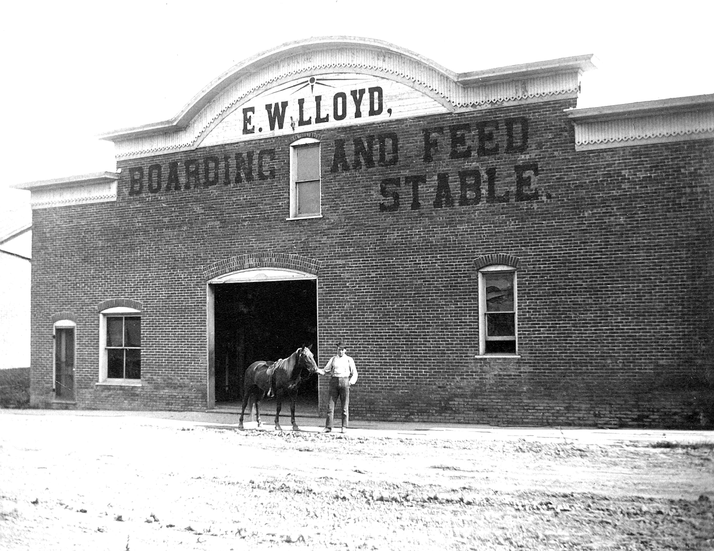 A black and white historic photo of a brick building with the sign 'E. W. Lloyd, Boarding and Feed Stable' on the facade. Two men stand at the entrance, one holding the bridle of a horse.