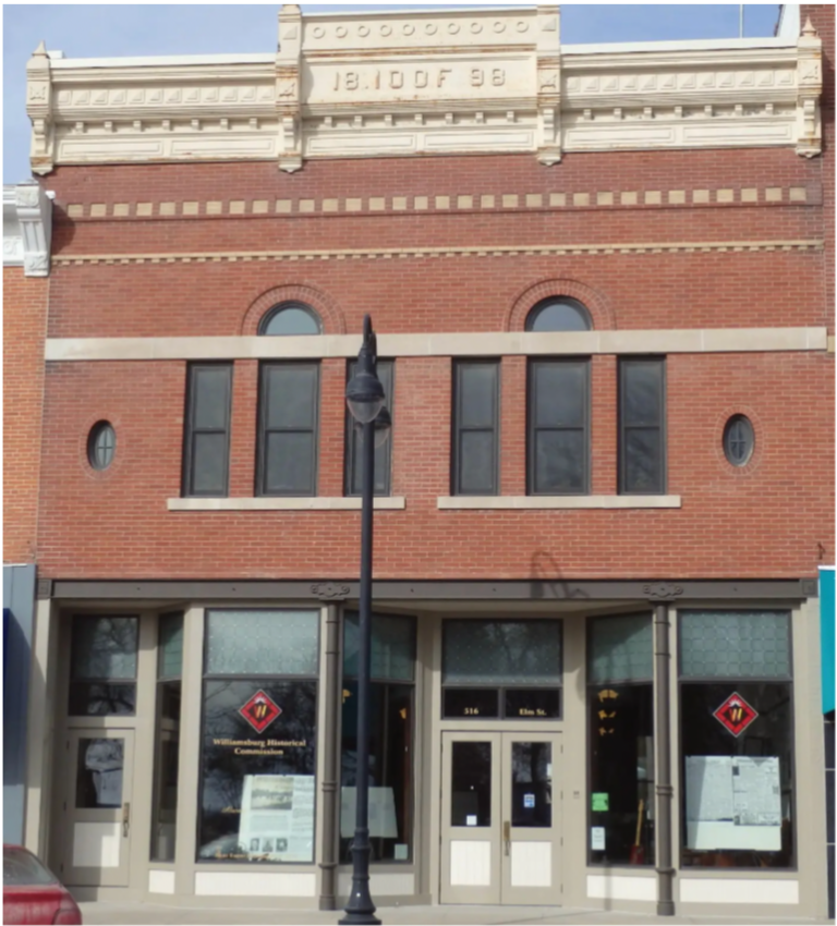 Street view of a brick building with three large windows on the second floor, a decorative cornice at the top, and a storefront on the ground floor with glass doors and windows, lit street lamp in front.