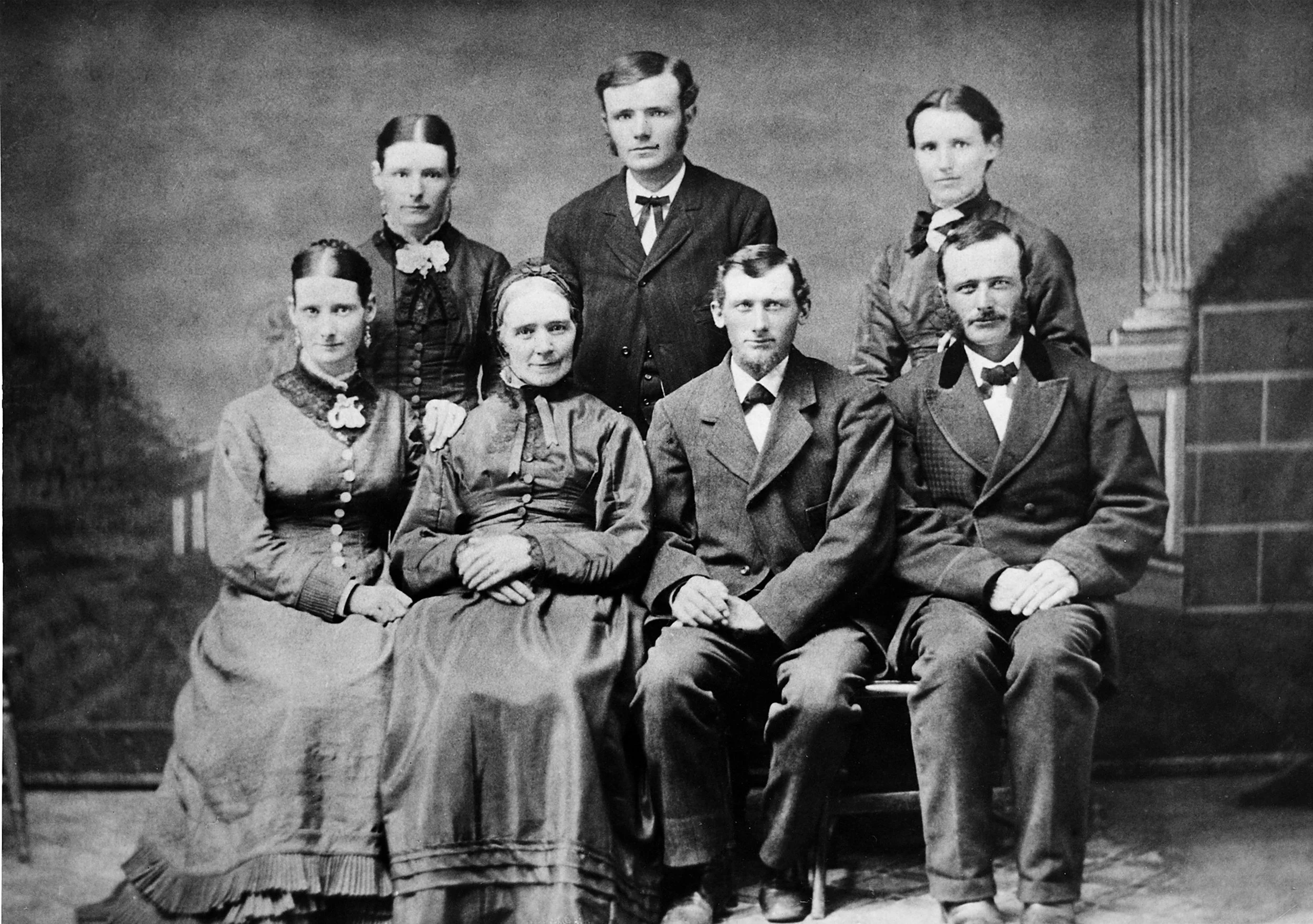 A black and white photograph of seven people, three men and four women, posing indoors with a plain background and classical architectural elements visible on the right. The women are seated in the front row, wearing dresses with high necklines and ornate details. The women have their hair tied back and appear to be dressed in 19th-century fashion. The men are standing in the back row, wearing suits with bow ties, with short hair. They all have serious expressions and are looking directly at the camera.