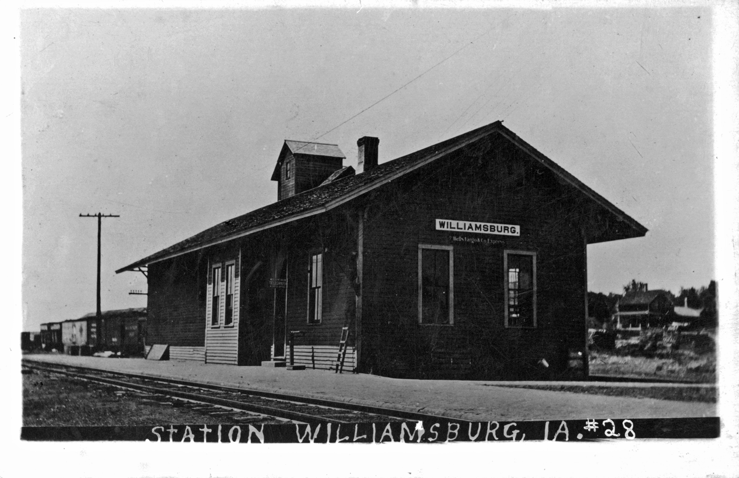 Black and white photo of a train station building with a sign that reads 'Williamsburg', a chimney, and a small tower on the roof. There are train tracks in the foreground and a train in the background. A telephone pole is visible on the left.