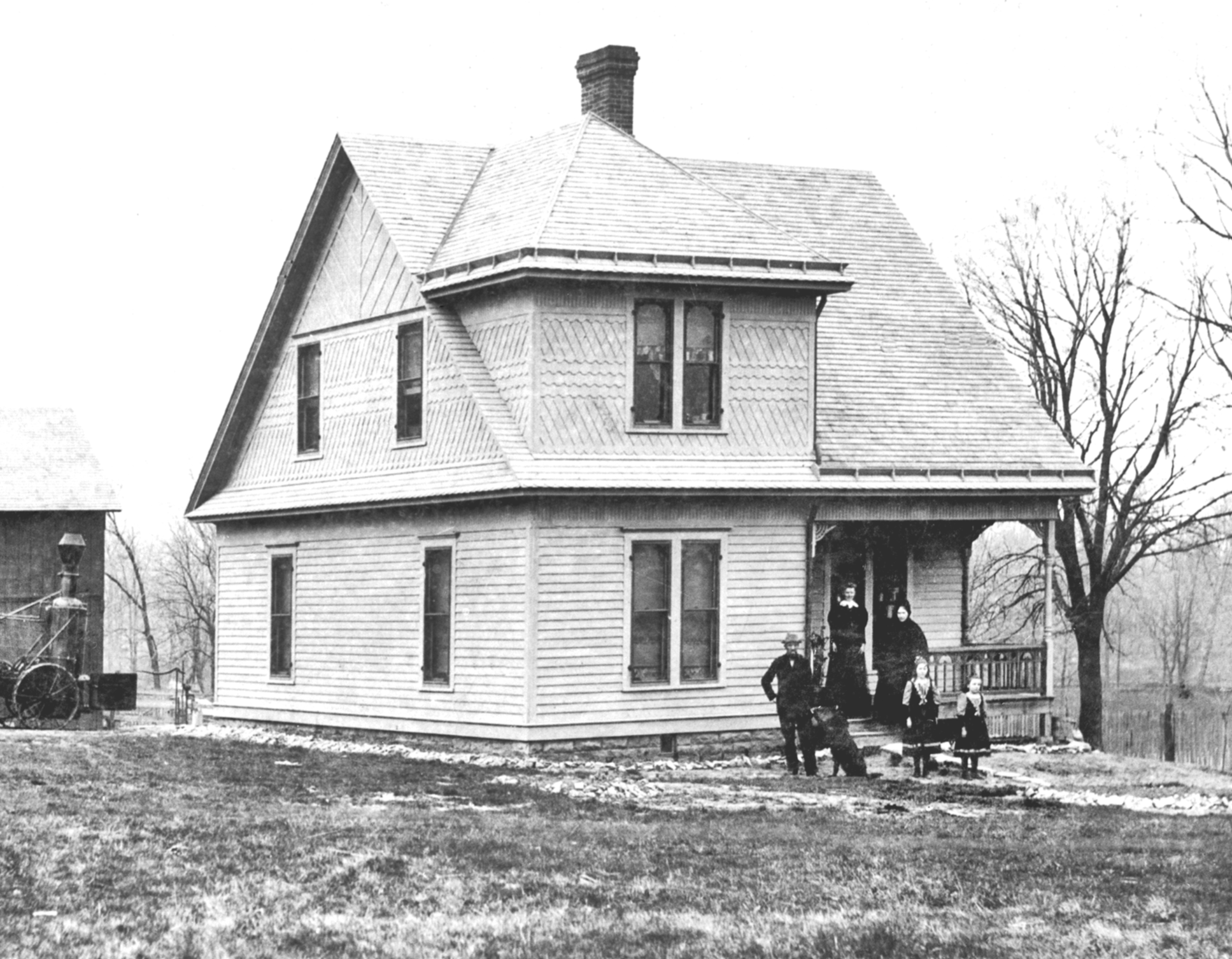 Black and white photo of a large, Victorian-style house with a steep roof, a chimney, and multiple windows. Several children and an adult stand in front of the house on a lawn. There are leafless trees and a fence in the background.