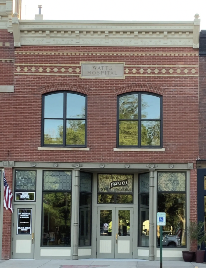 Front view of a historic brick building with a sign that reads "Watt's Hospital" at the top. The ground floor features a drug store with glass doors and windows, and a sign that says "Drug Co." The building has two large arched windows on the second floor, and an American flag is visible on the left side.