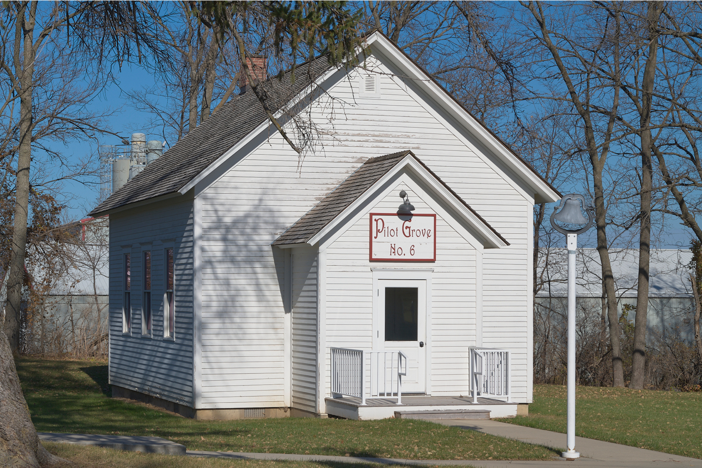 White wooden building with a sign that reads 'Pilot Grove No. 6,' a small porch with railing, and a bell post nearby. Trees with bare branches surround the building, and there is a clear blue sky.