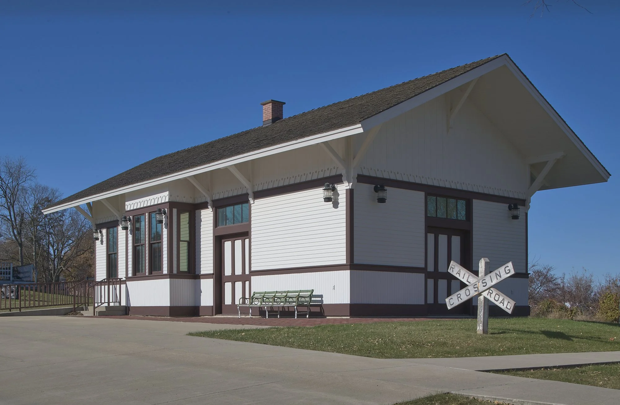Historic white train depot building with dark trim, large windows, and a railroad crossing sign in front. The building sits beside a paved walkway with grass and trees under a clear blue sky.