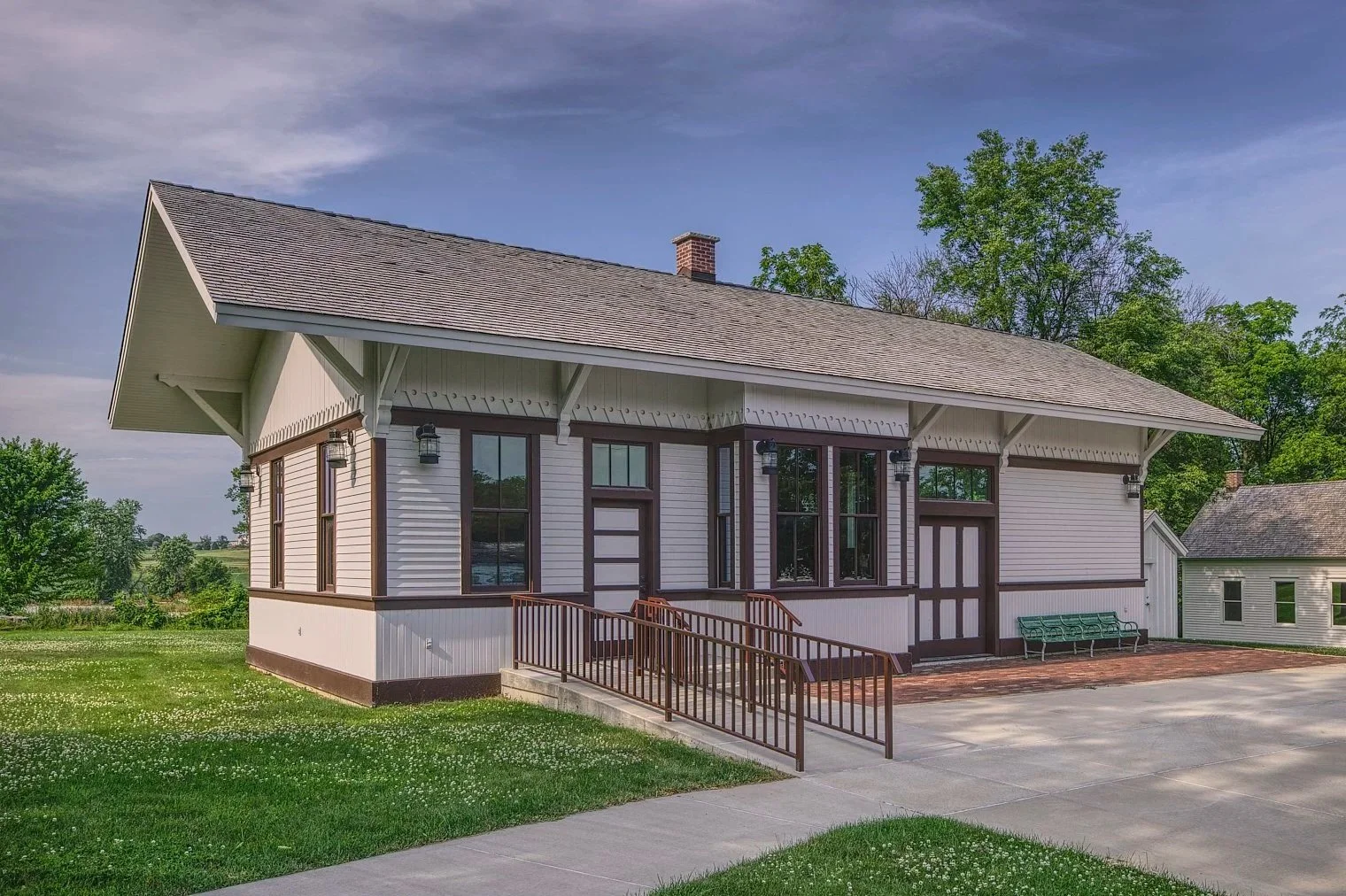 A small, white, wooden house with dark brown trim, a front porch with a ramp, large windows, and outdoor wall-mounted lights, surrounded by green grass and trees under a partly cloudy sky.