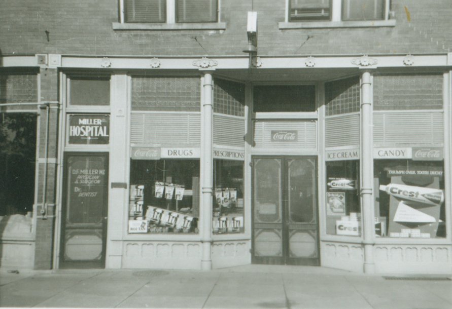 An old black and white photo of a small storefront with signs for a hospital, Coca-Cola, drugs, prescriptions, ice cream, and candy. The display window shows products inside, and the entrance has glass doors.