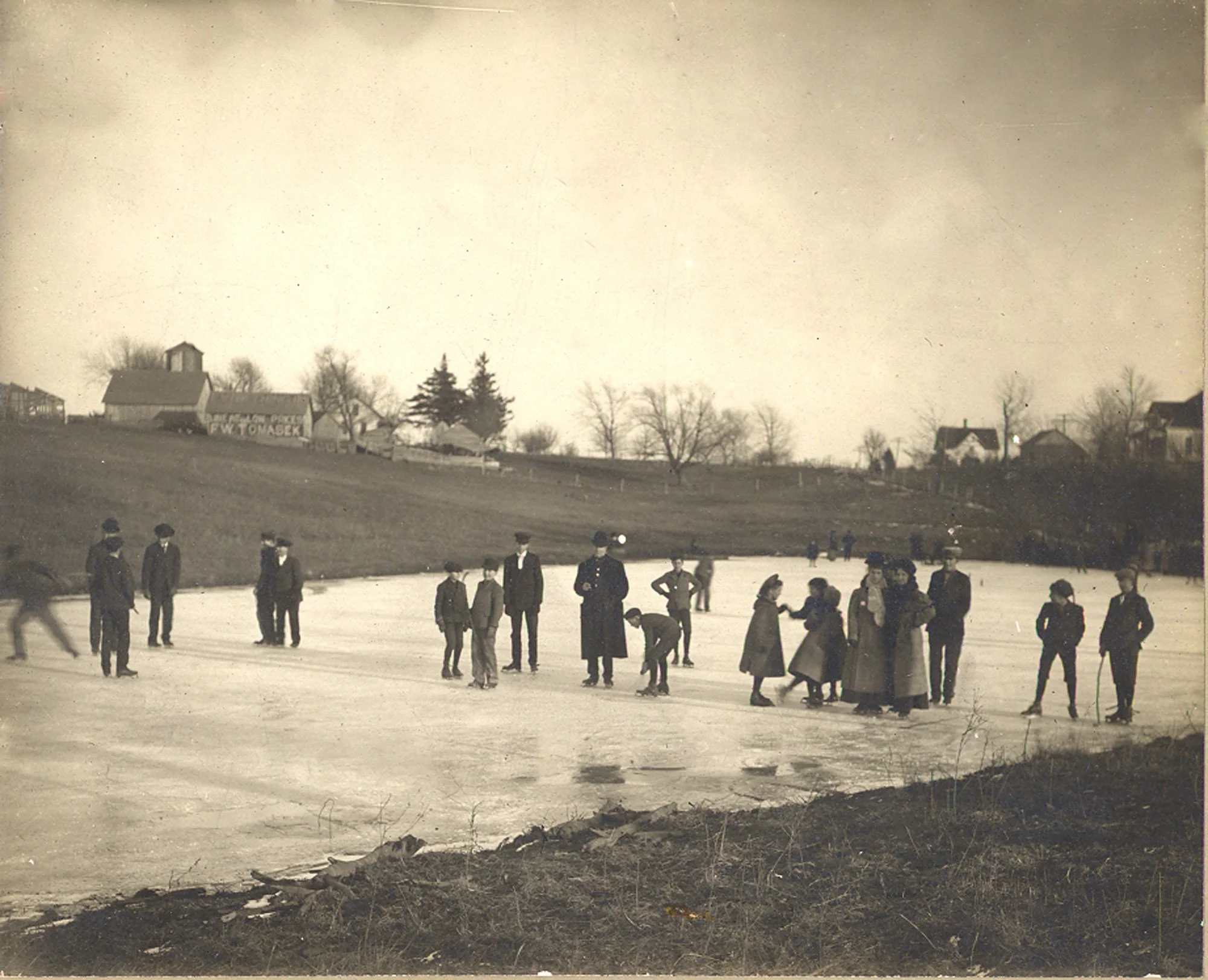 A historical black-and-white photograph of children and adults standing in an open field with houses and trees in the background, likely from the early 20th century.