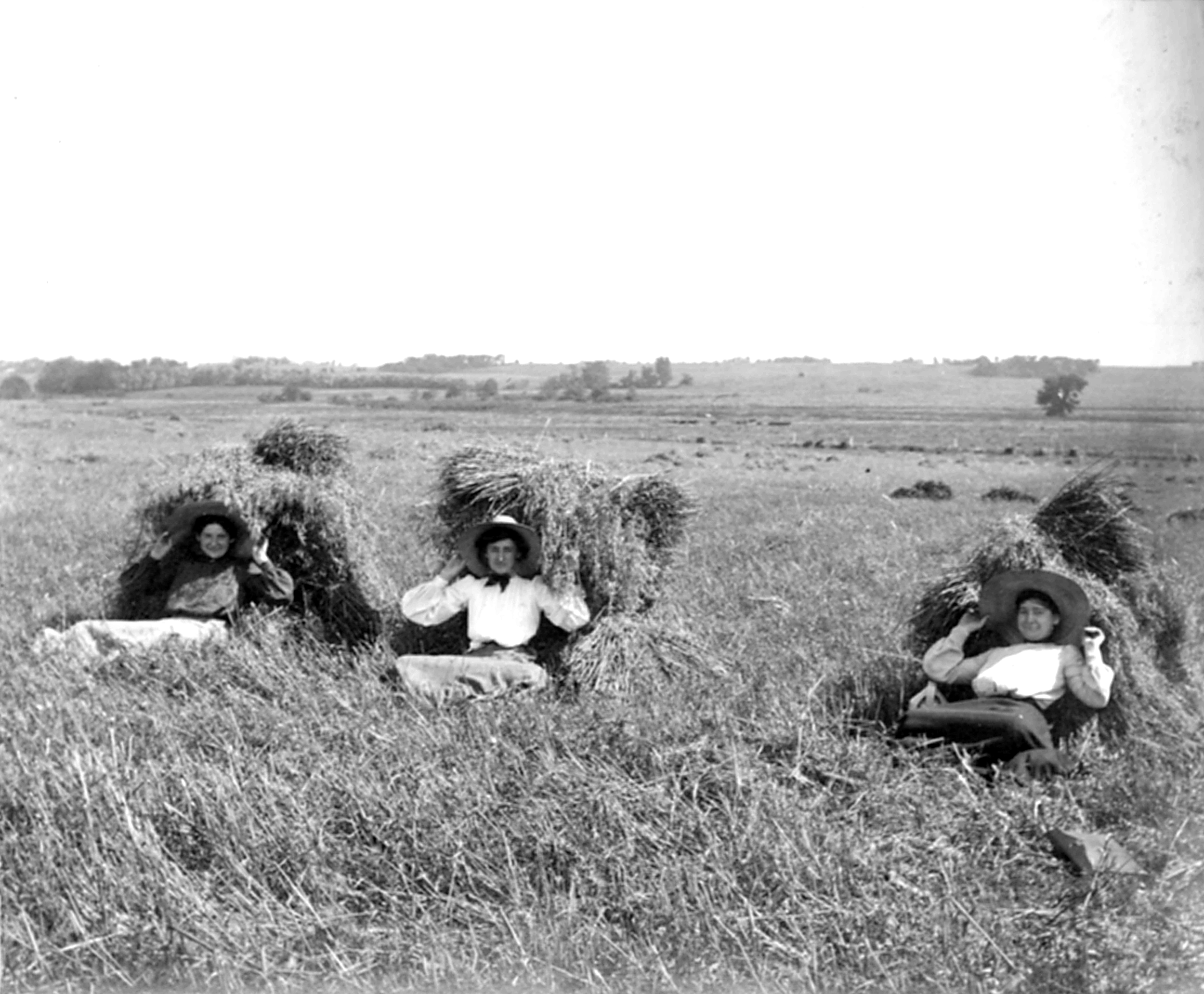 Black and white photo of a rural landscape with four women sitting and lying in the tall grass in the foreground, showing a wide open field with a few trees and distant hills in the background.