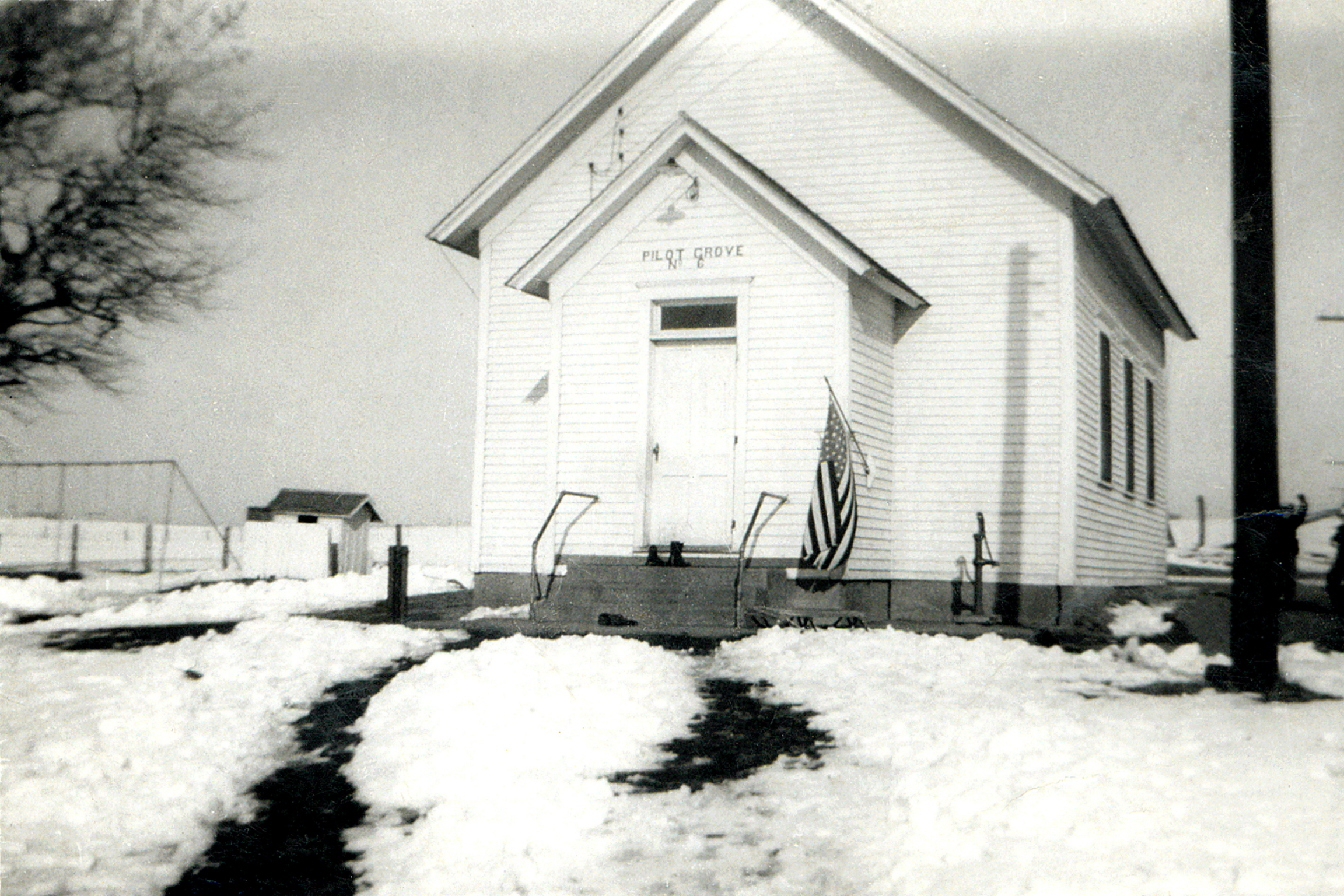 A white wooden building with a gabled roof, labeled 'Pilot Grove No. 6,' surrounded by a fence and snow-covered ground, with an American flag hanging outside.