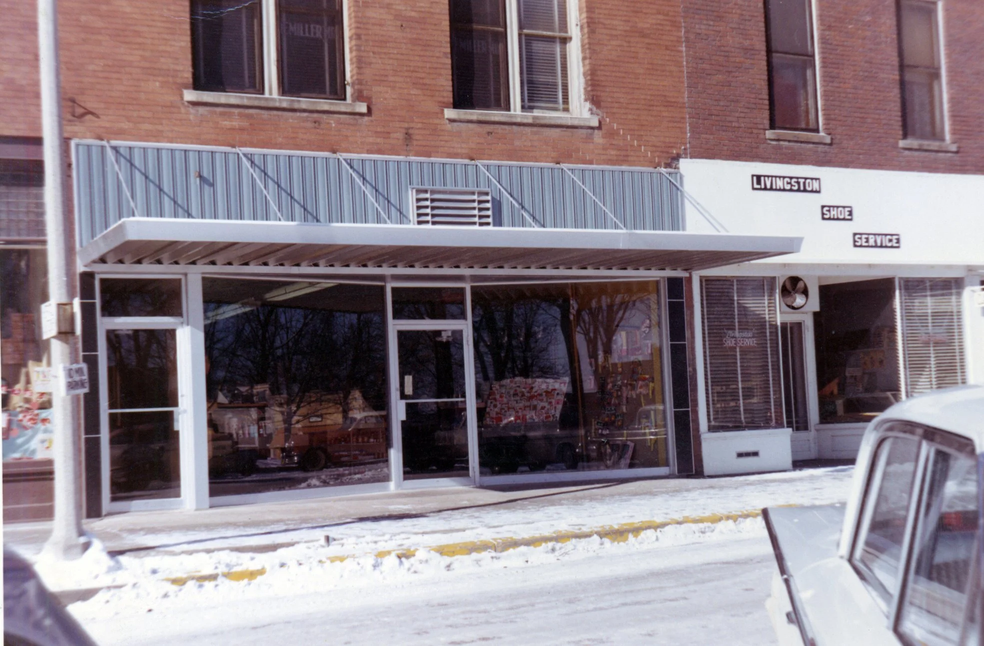 Street view of a shoe repair store named 'Livingston Shoe Service' with large glass windows, a striped awning, and brick building facade, snow on the sidewalk, and a sidewalk sign on the left.