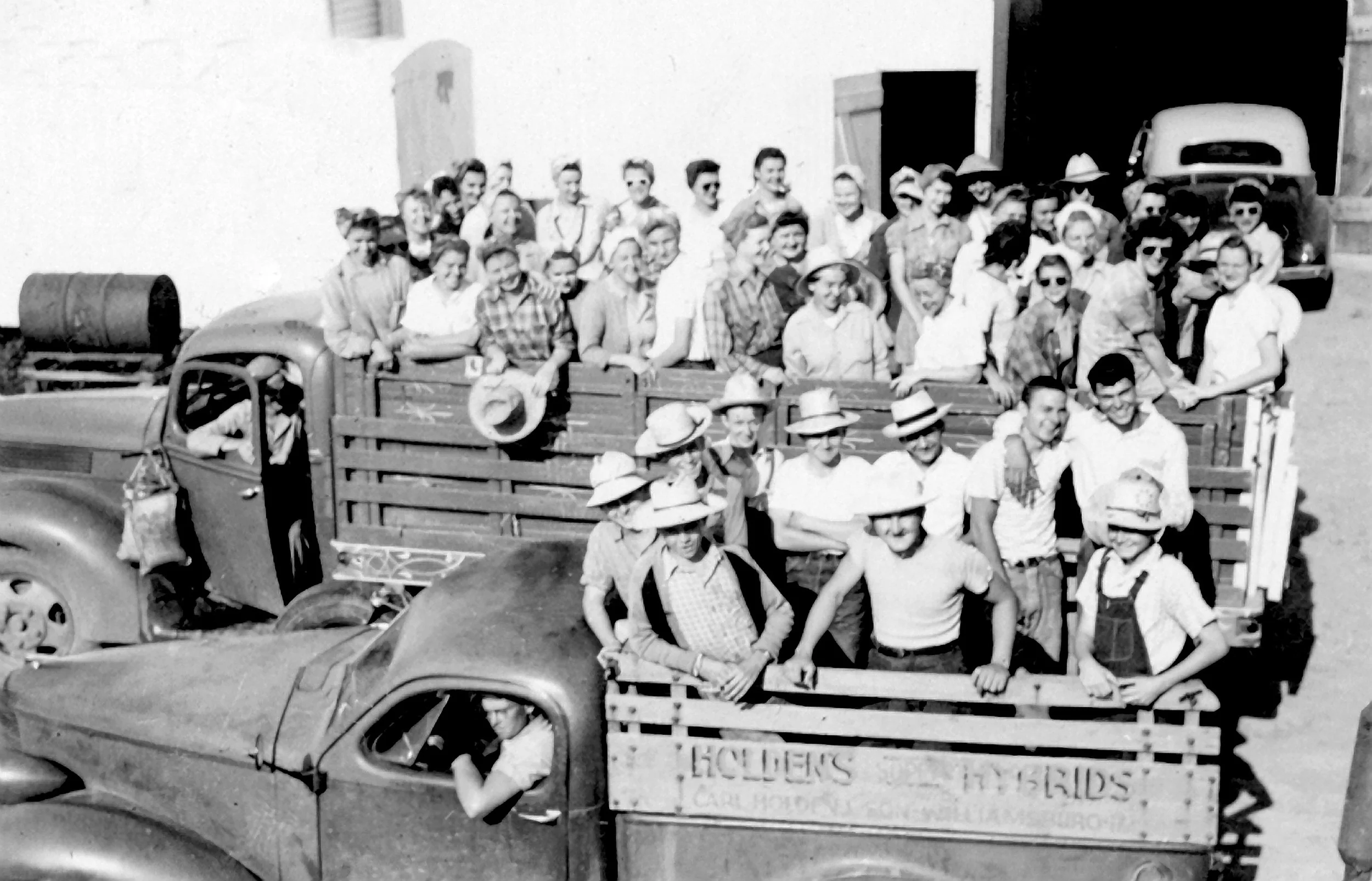 Black and white photo of a large group of people, including men, women, and children, gathered on a truck, possibly during a parade or community event, with some individuals wearing hats and sunglasses. The truck has a sign on the side reading, 'HOLLER'S,' and appears to be parked outside a building or warehouse.