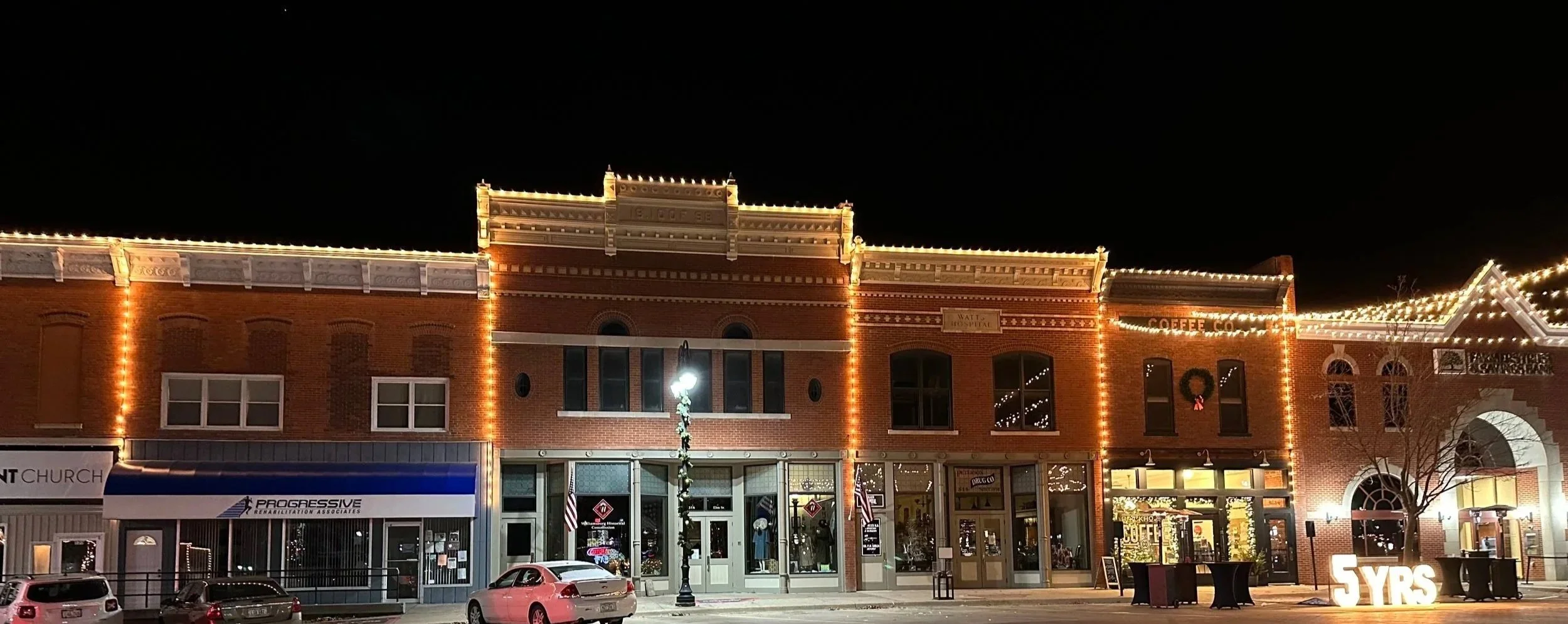 Night view of a decorated brick building with string lights outlining the roof and windows, featuring shops and a streetlamp in front.