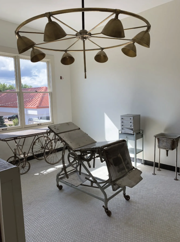 Museum room with vintage hospital equipment, including an adjustable hospital bed, a wheelchair, and IV stands, with a window showing a red-roofed building outside and a modern light fixture on the ceiling.