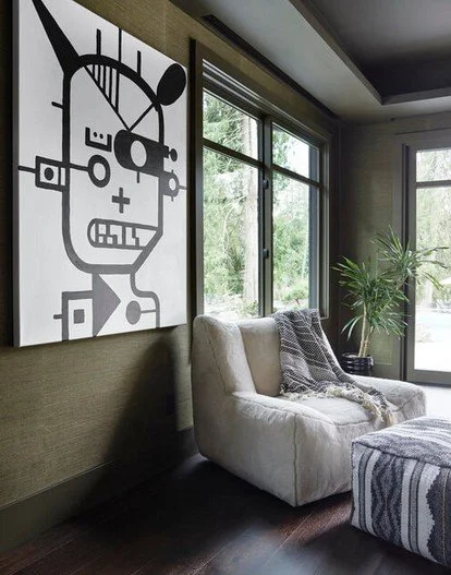 Living room with a white armchair, a large abstract black-and-white artwork on the wall, a tall green plant near windows, and a patterned ottoman.